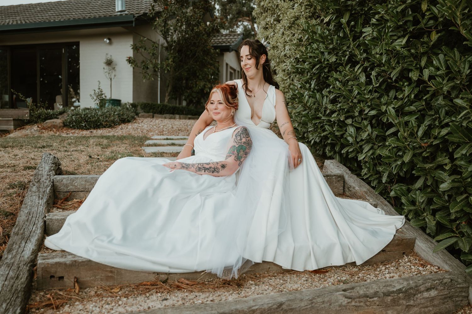 Wedding couple in white dresses pose together on rustic outdoor grounds.