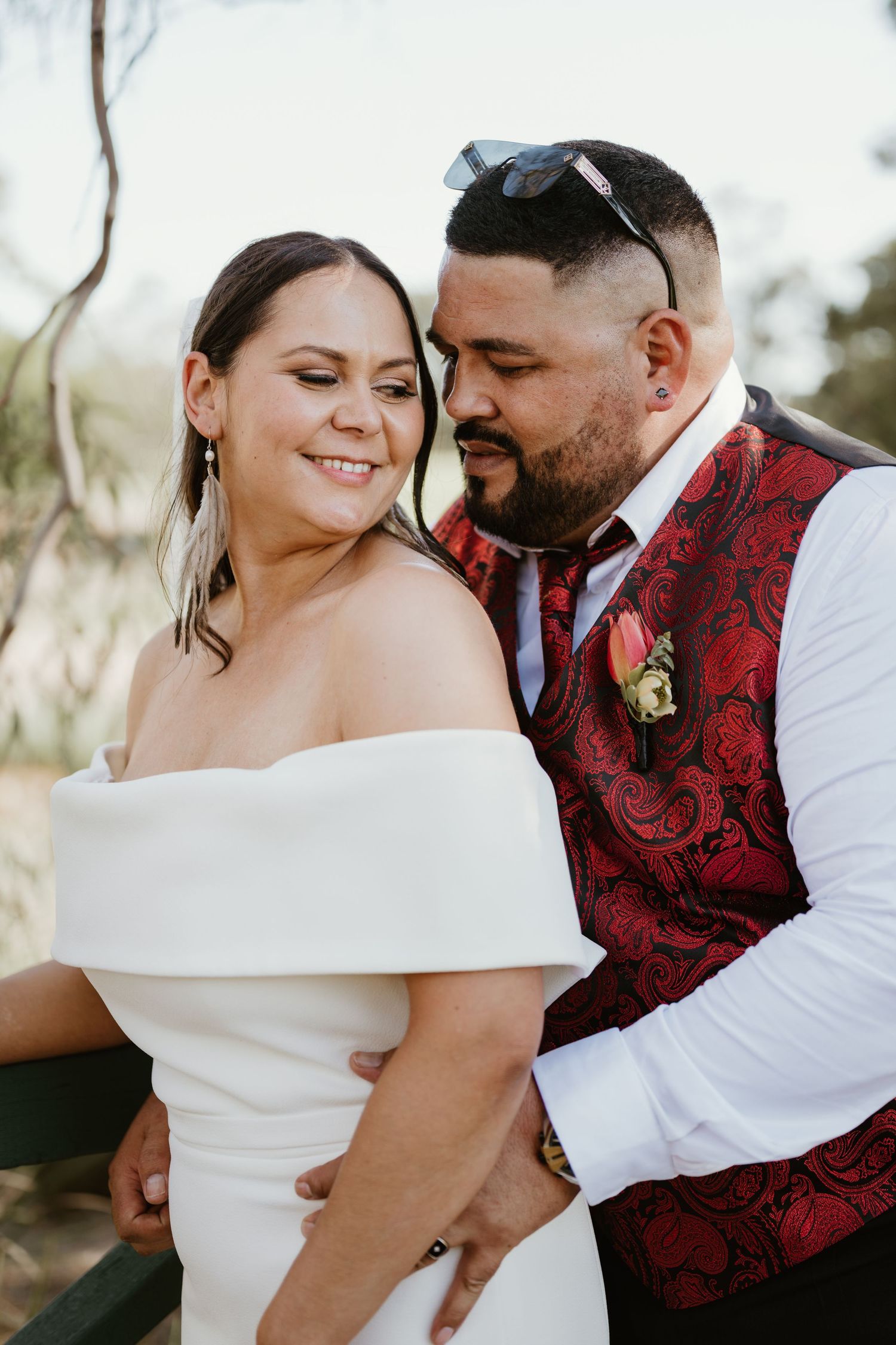 A romantic wedding moment captured outdoors with a bride in white and groom wearing a red paisley vest.