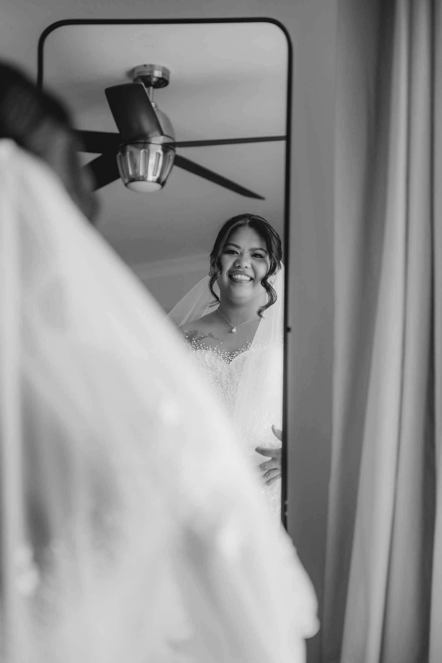 Black and white reflection in mirror shows a joyful bride in white dress with a ceiling fan visible above.