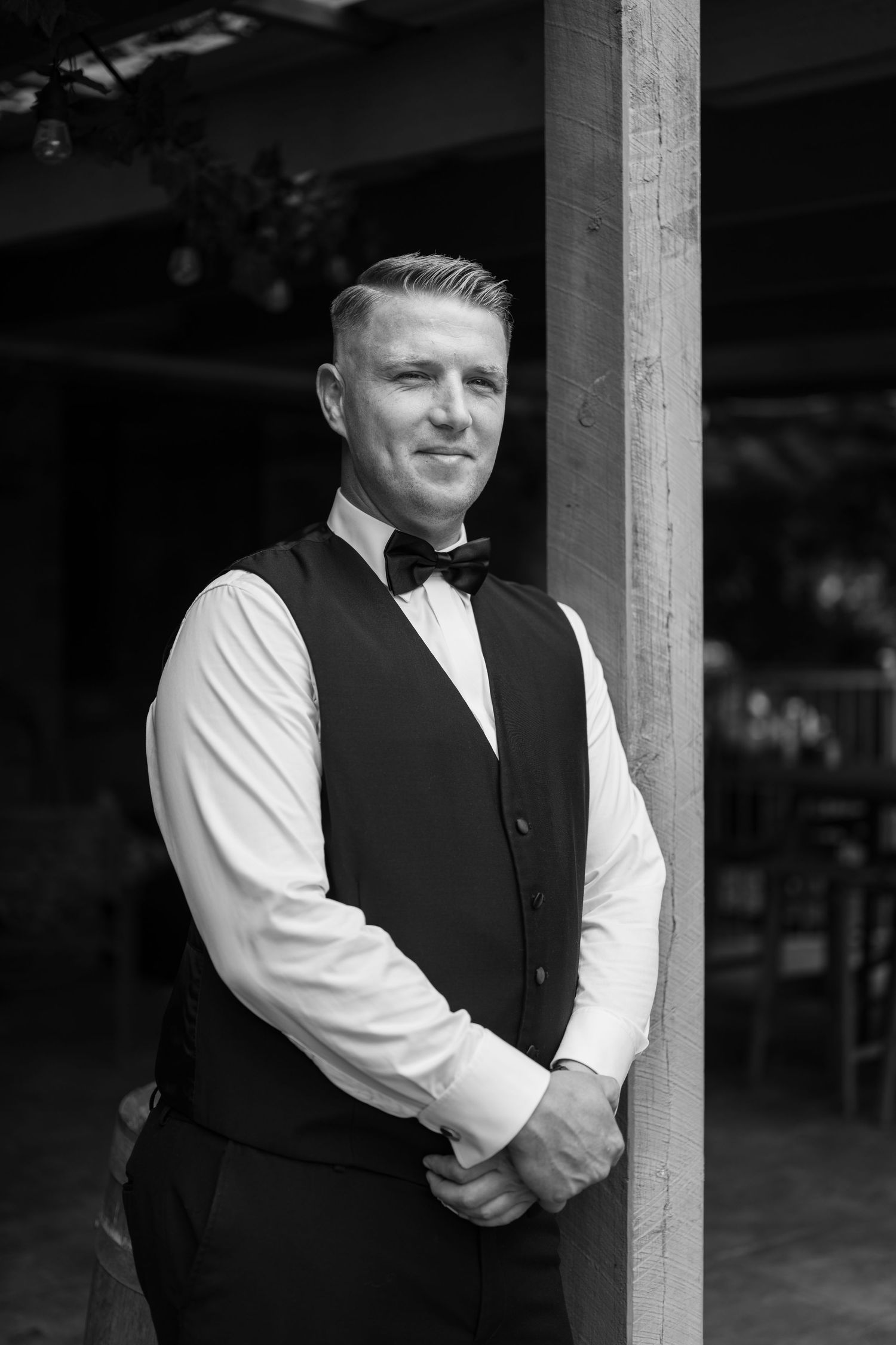 Person in formal vest and white shirt poses against rustic wooden post in black and white.