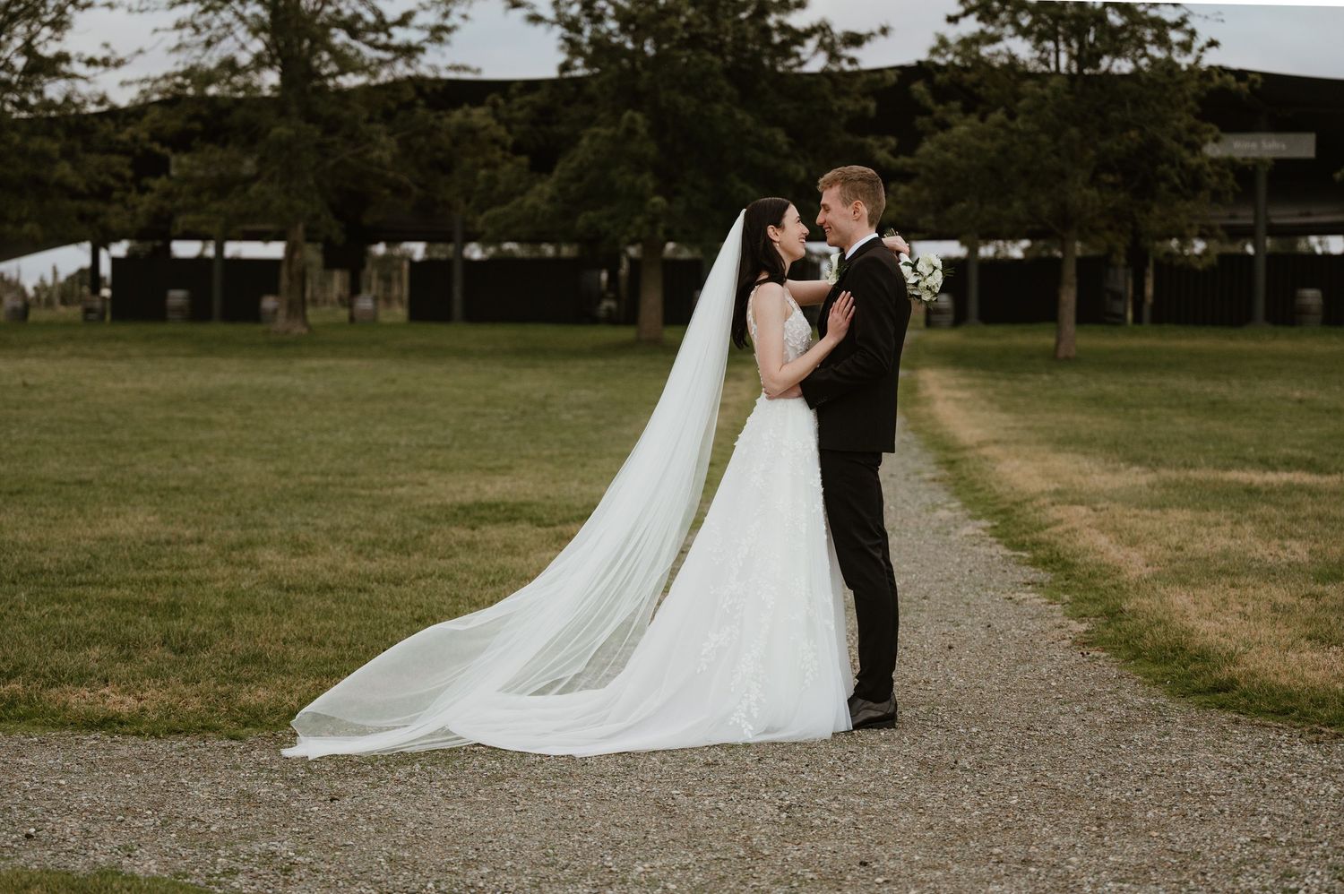 Wide shot of newlyweds walking together on a rustic gravel path surrounded by green fields.