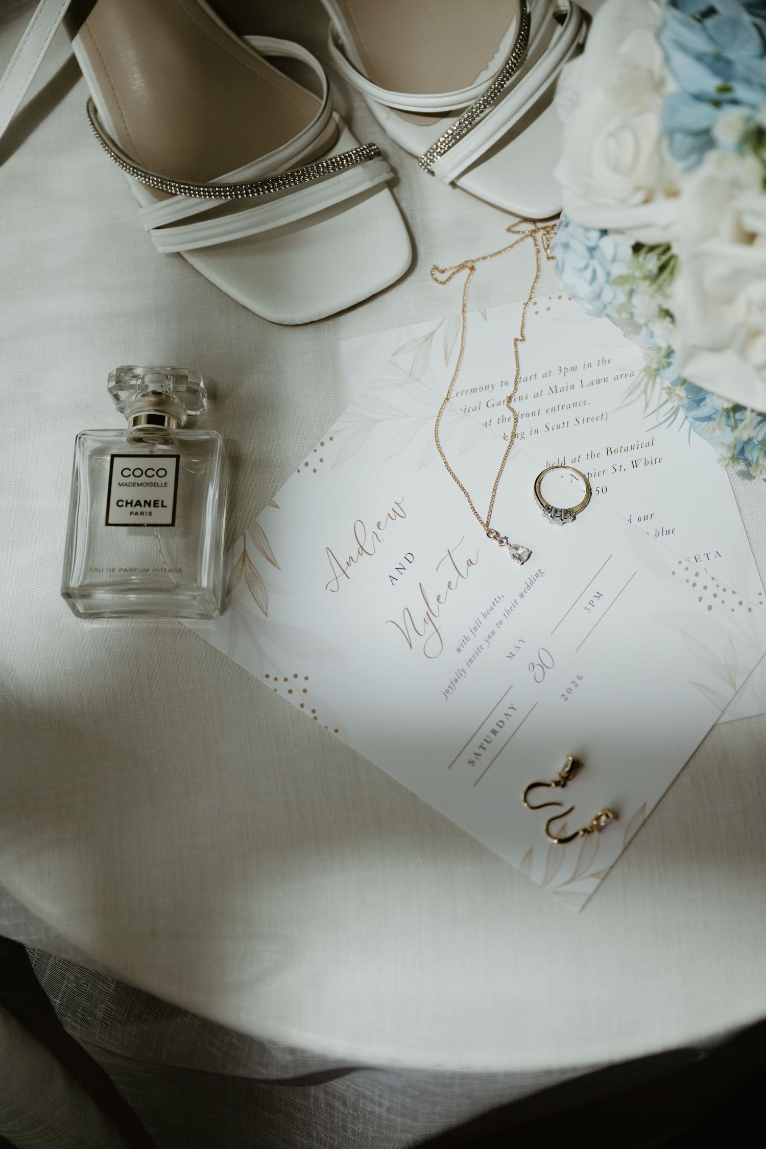 Wedding flat lay featuring Chanel Coco perfume, white heels, gold jewelry, floral invitation, and blue roses.