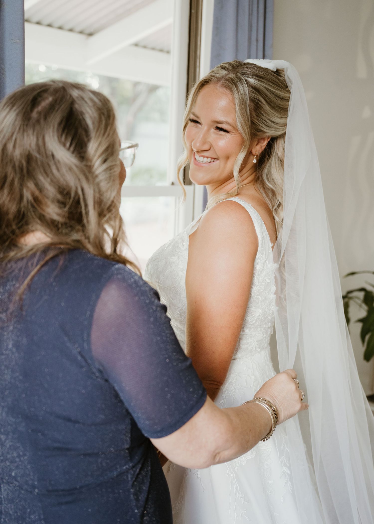 A smiling bride in a white gown and veil being helped into her dress by a woman in navy blue.