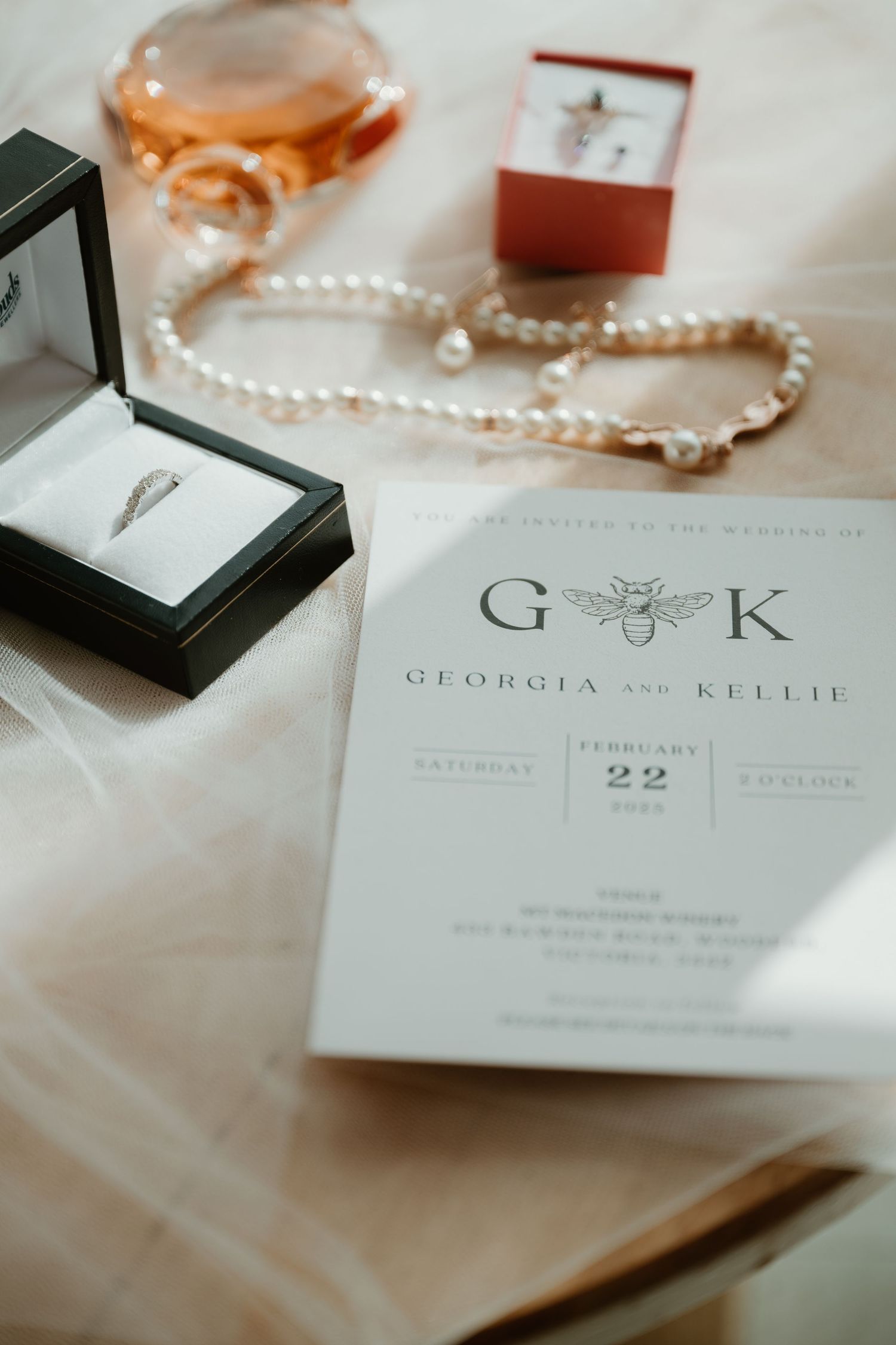 Elegant white wedding invitation suite with a bee logo and delicate pearl details on a light fabric background.
