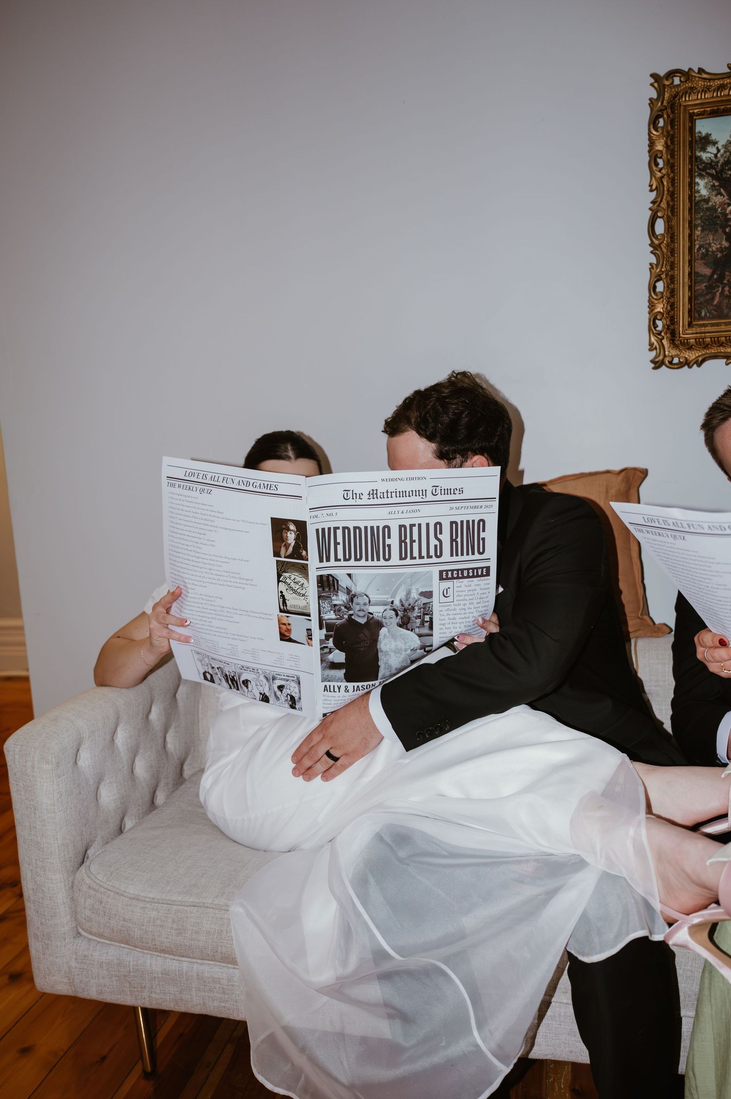 People sitting on a white tufted couch reading newspapers together in a staged wedding photo series.