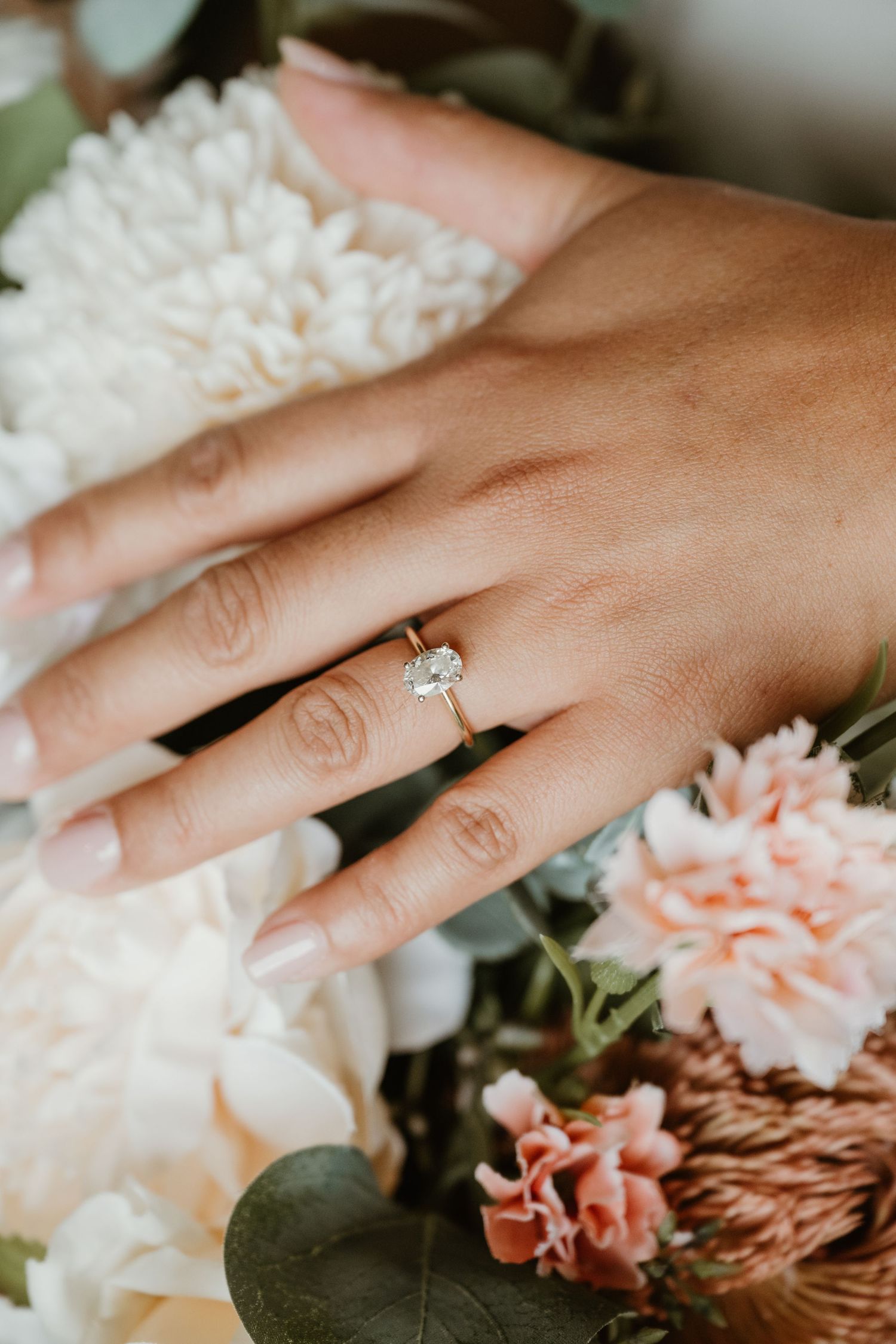 A hand wearing an elegant diamond engagement ring rests among delicate pink and white flowers and petals.