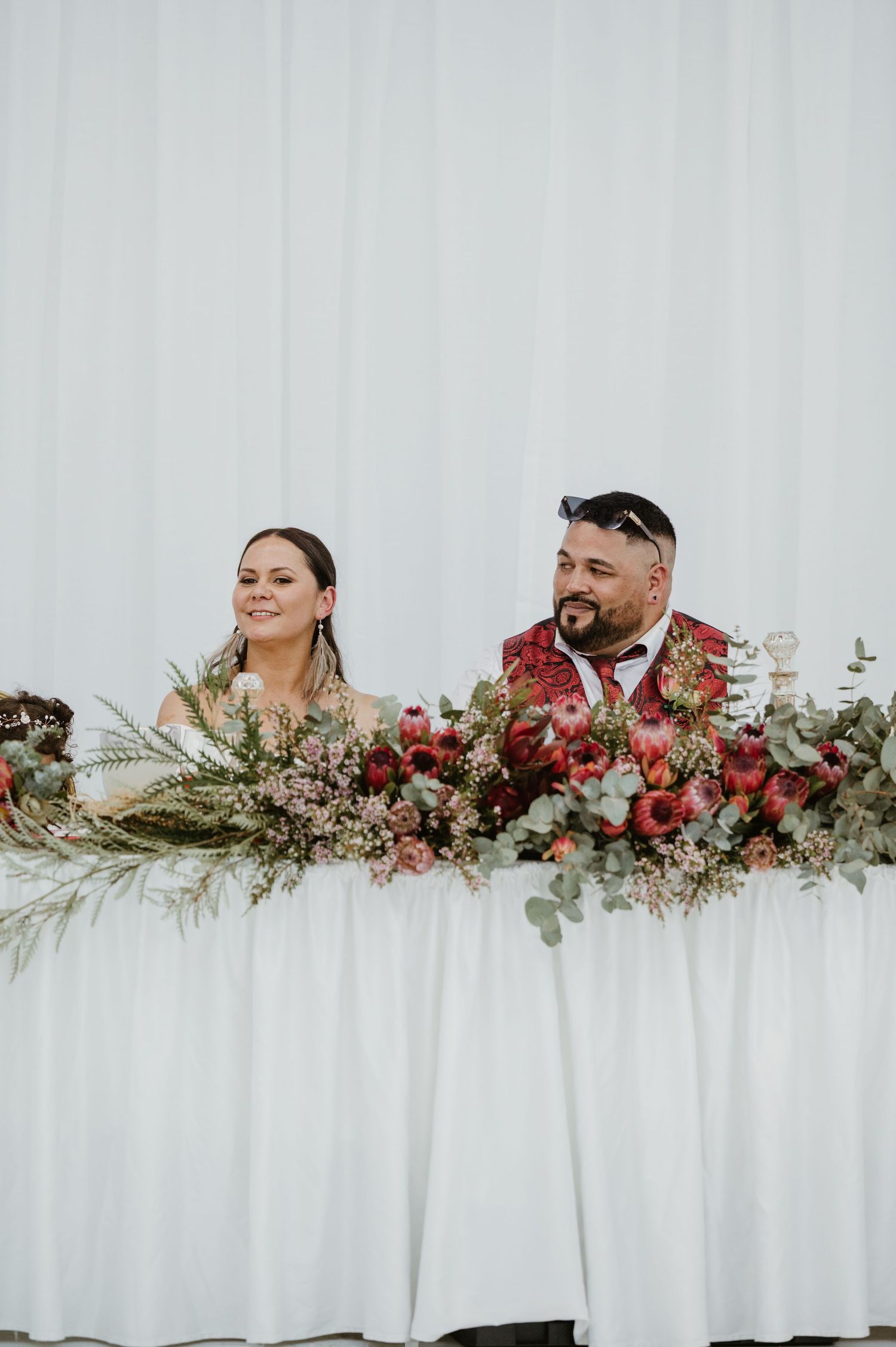 Wedding party members sit behind an elegant floral table arrangement with red roses and greenery against a white backdrop.