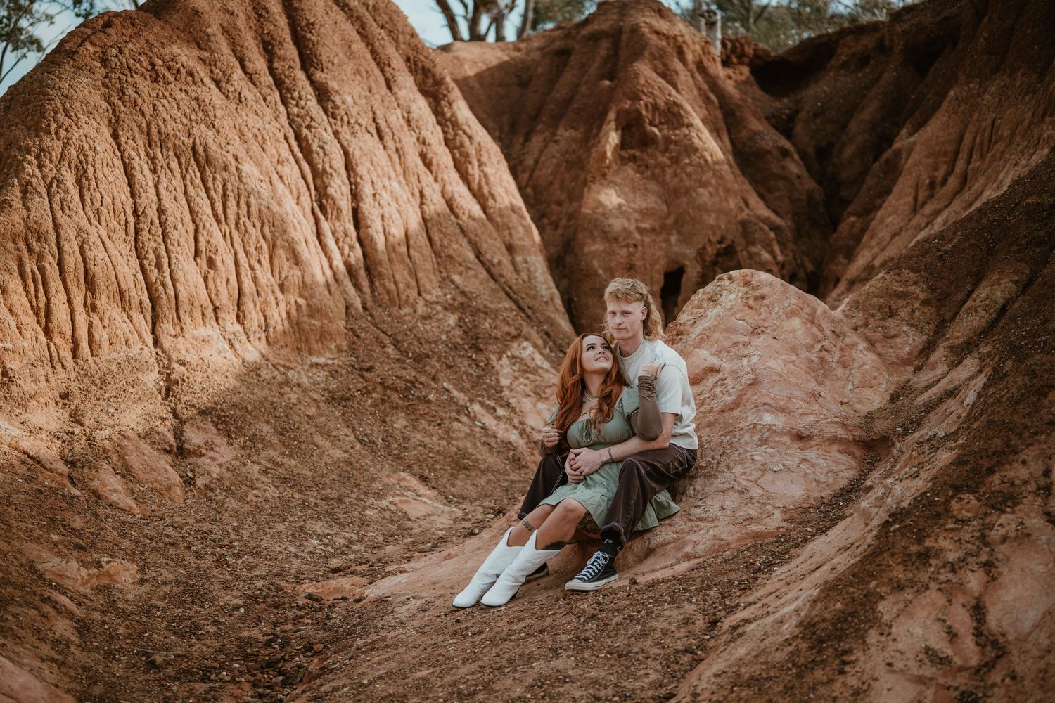 A couple sits together on sandy terrain during a romantic outdoor photoshoot.