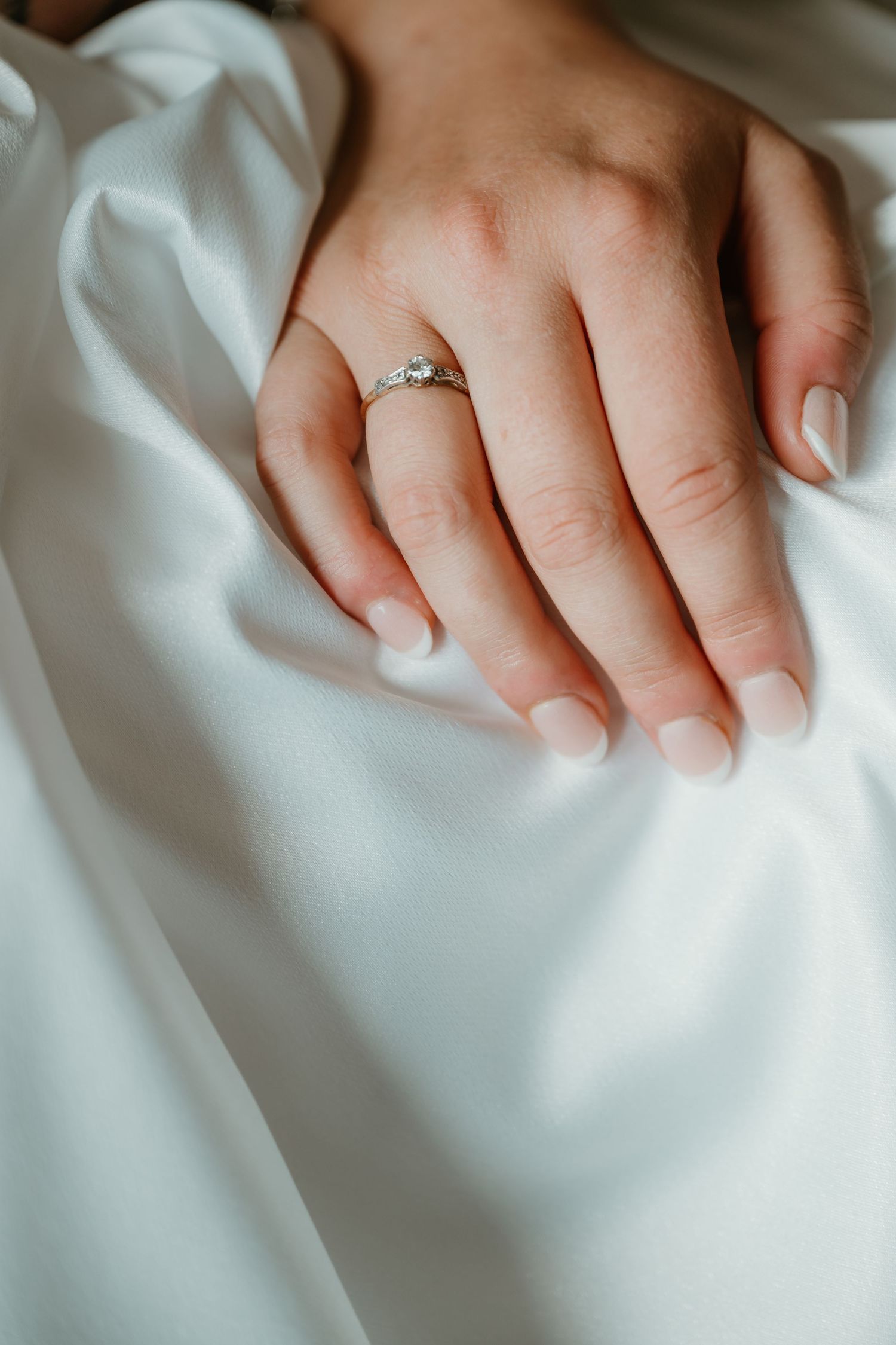 Close-up detail of hands resting on white wedding dress fabric with engagement ring.