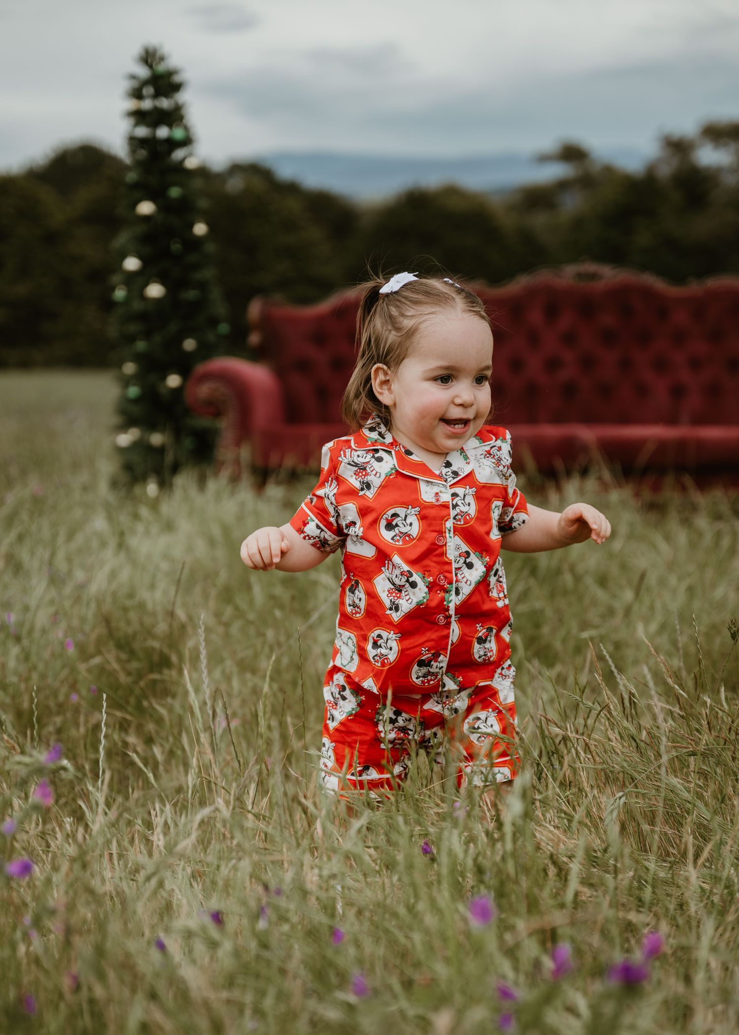 Toddler girl in red floral romper standing in a grassy field with a Christmas tree and red sofa backdrop.