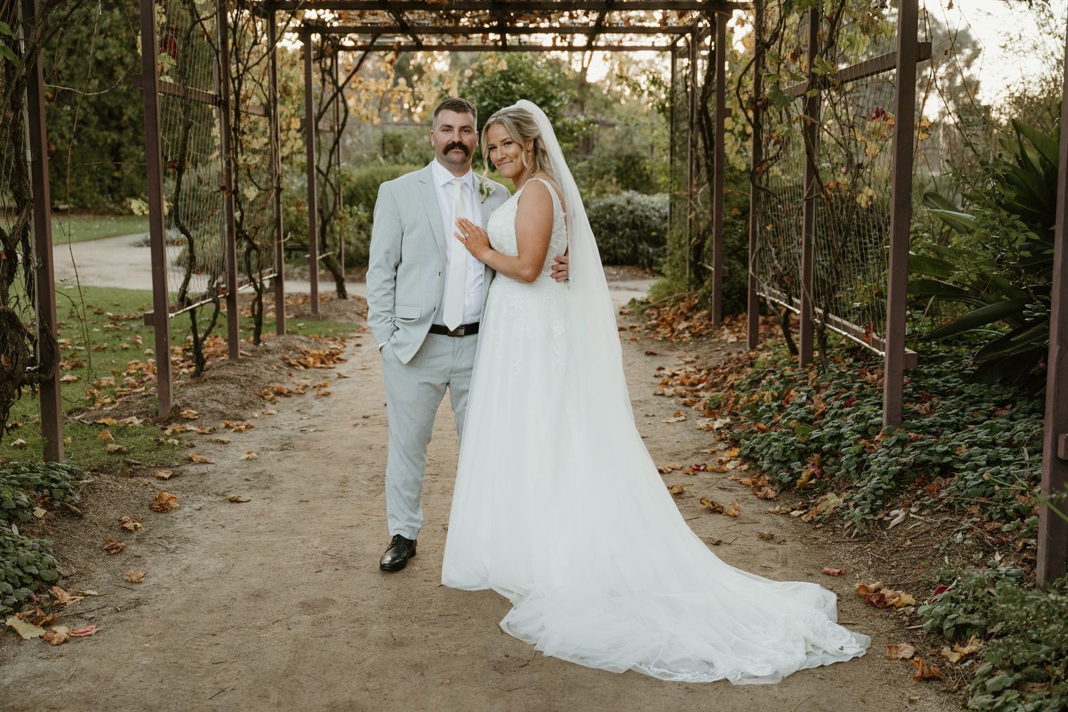 Bride and groom walk hand in hand along a garden path under a rustic pergola, surrounded by lush greenery.