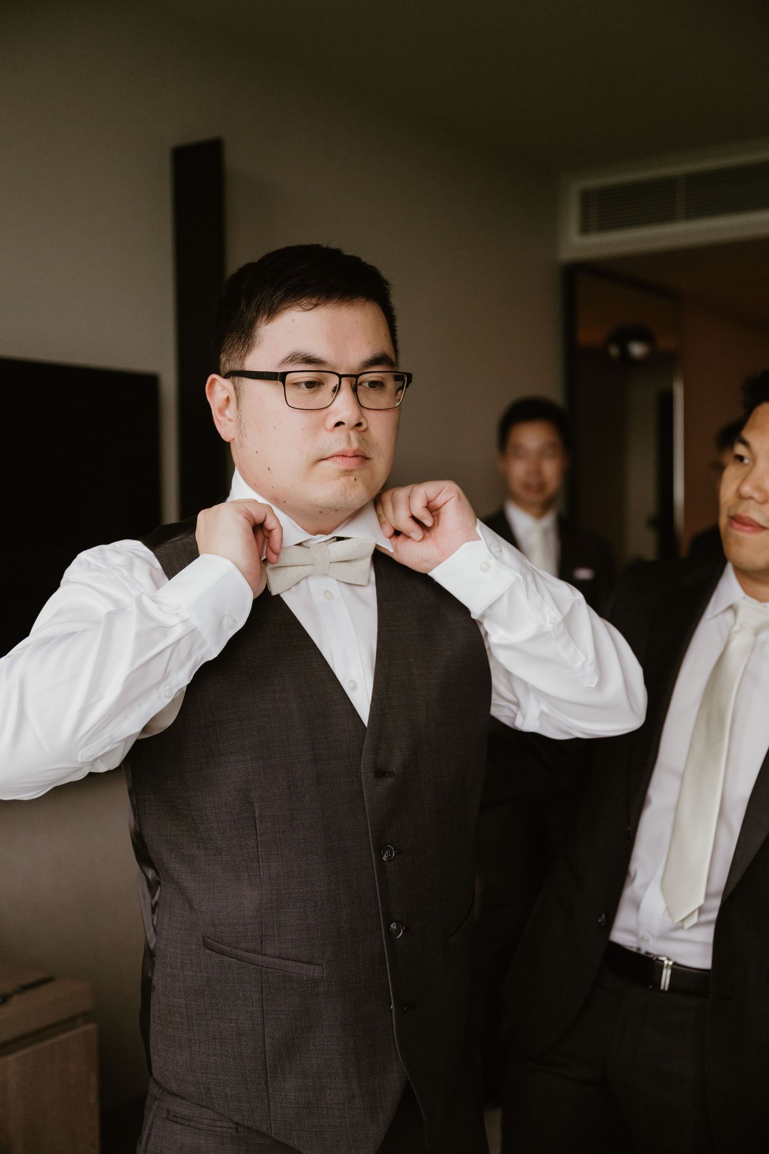 A person in a dark vest and white shirt adjusts their glasses while posing in a dimly lit room.