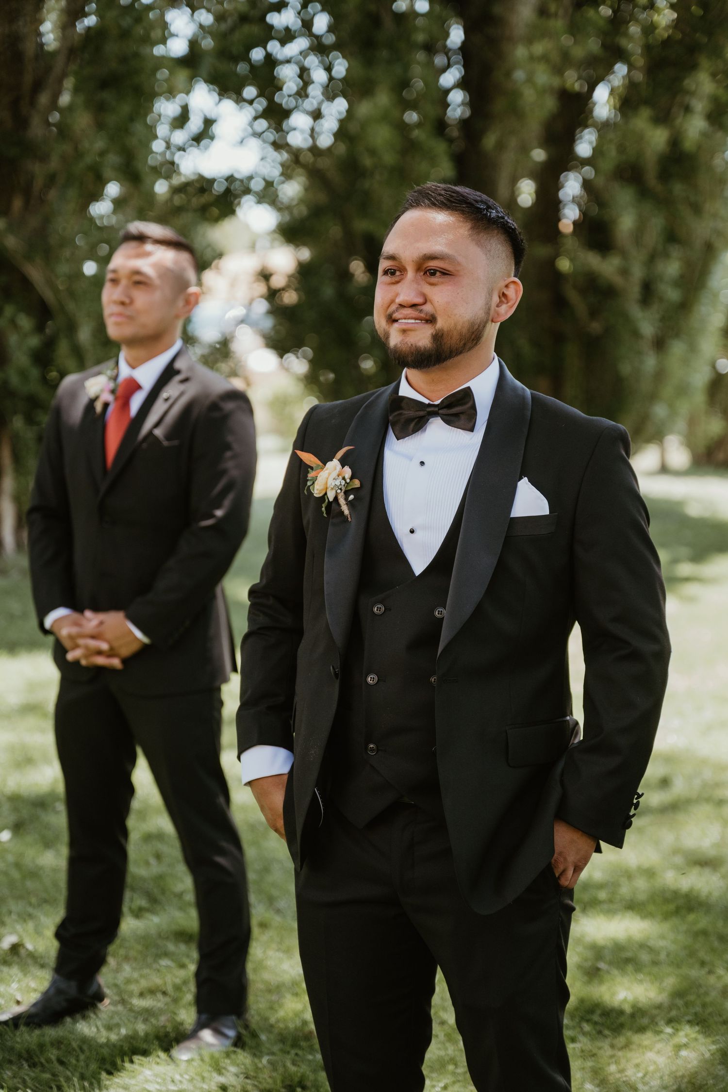 Groomsmen in classic black tuxedos and bowties stand together on grass during an outdoor wedding ceremony.