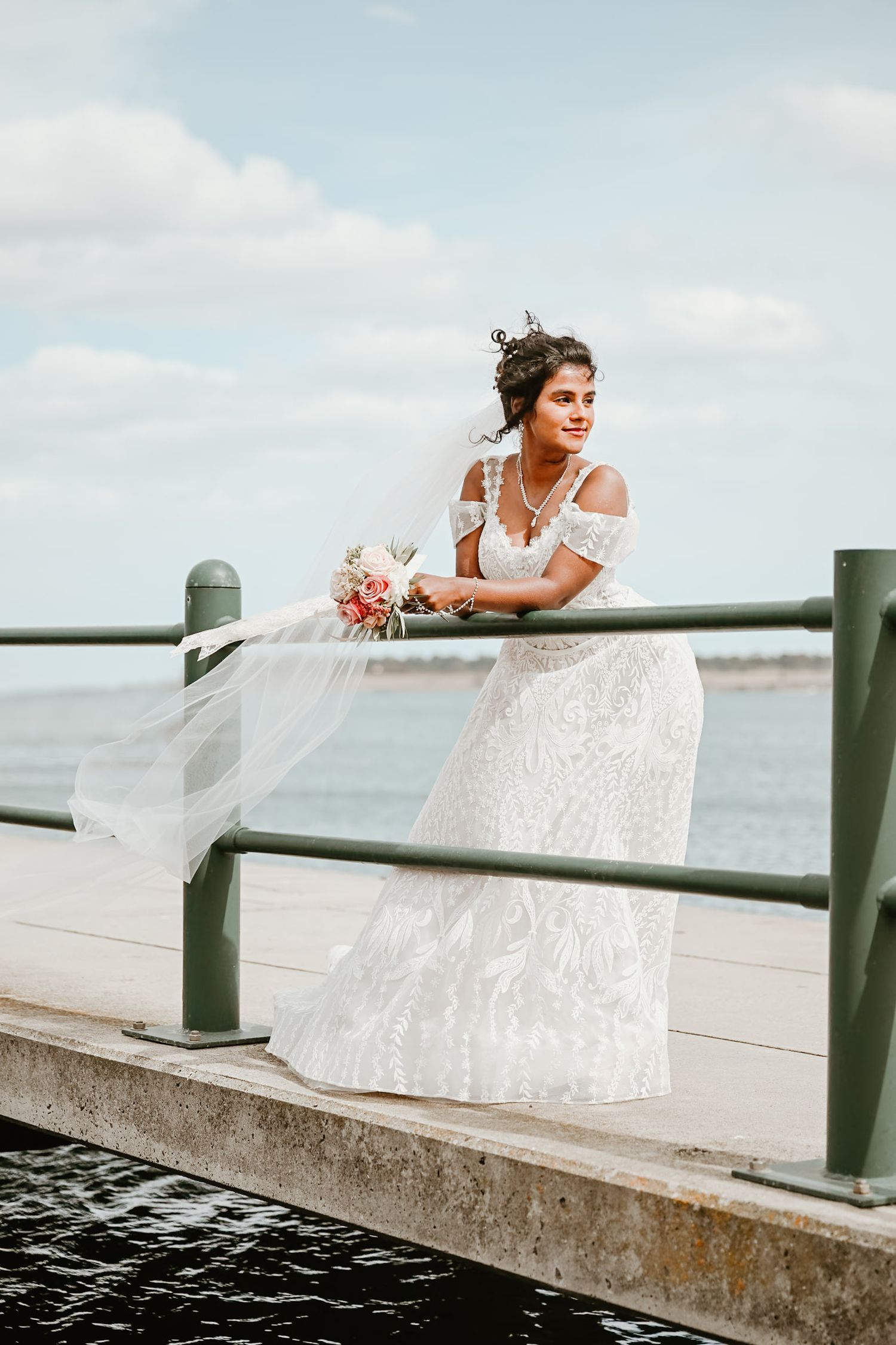 Bride in lace dress poses playfully on waterfront railing.
