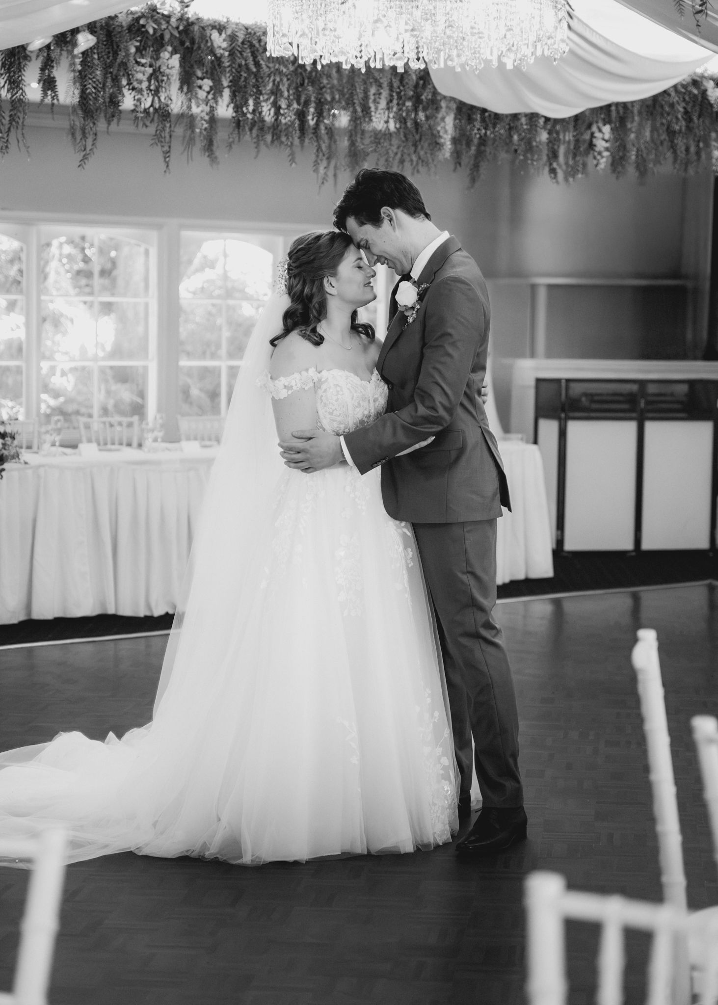Black and white photo of bride and groom sharing first dance at elegant wedding reception with floral chandelier.