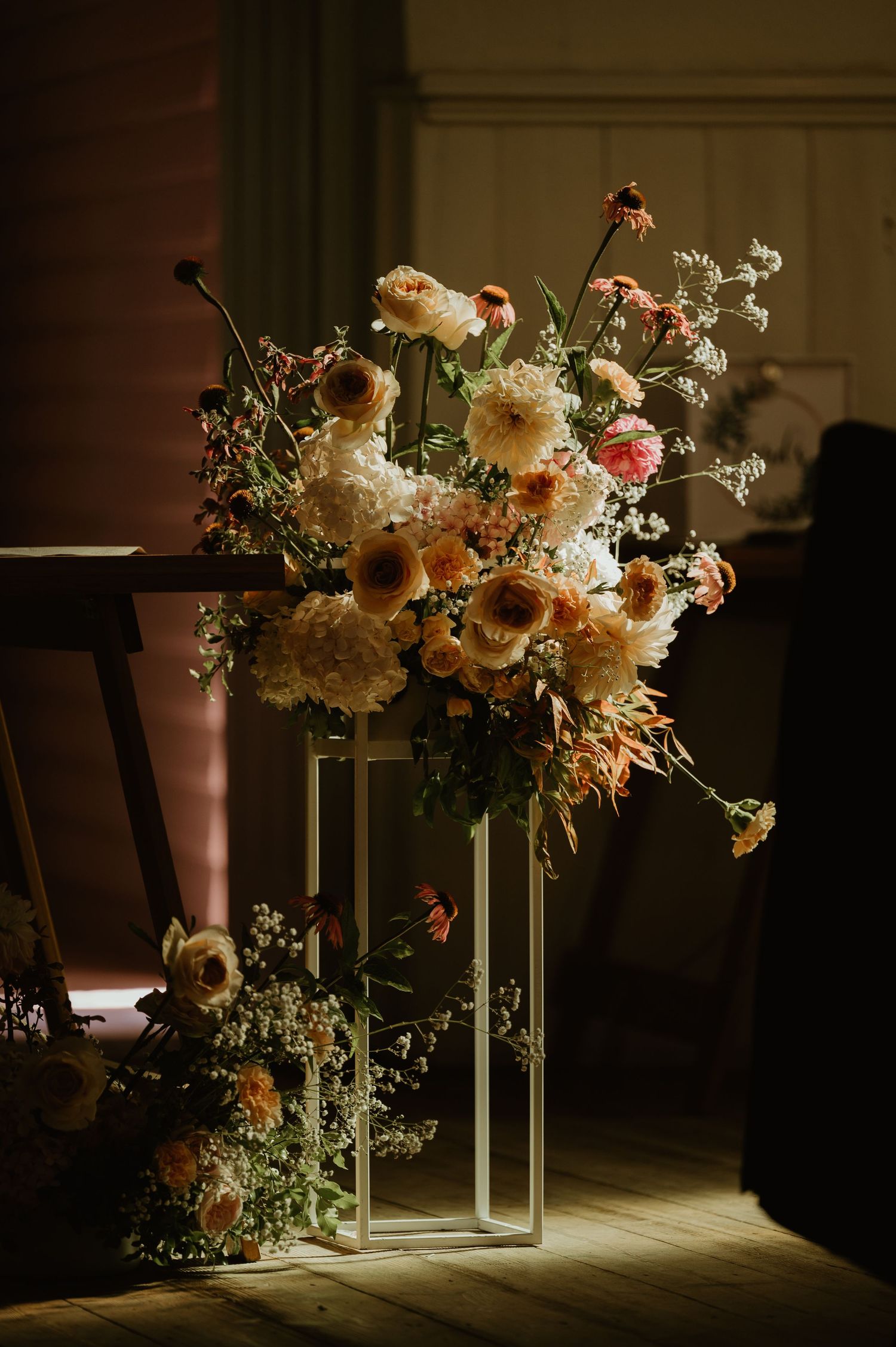 A moody floral arrangement with white and peach blooms in a modern geometric vase sits in dramatic lighting.