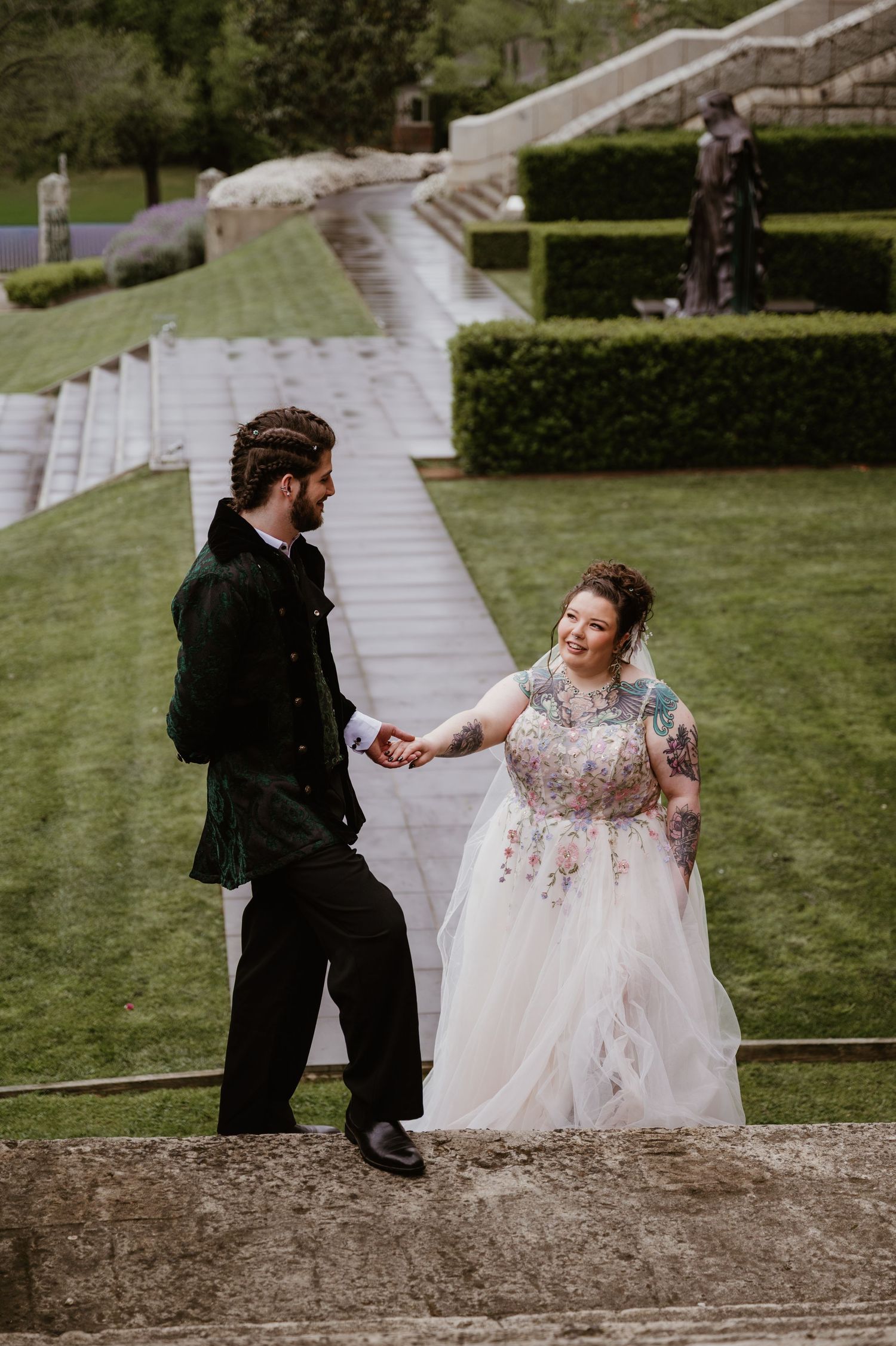 A couple in wedding attire dances and twirls on stone steps surrounded by a lush green lawn and garden setting.