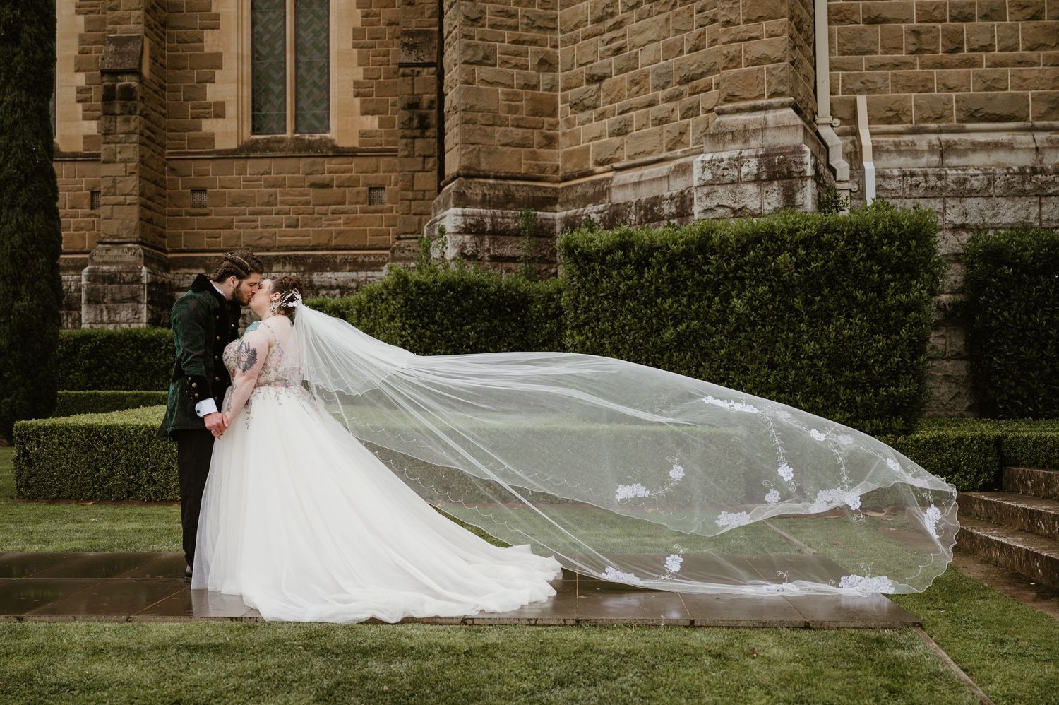 A flowing white wedding veil blows dramatically in the wind against a stone church building backdrop.