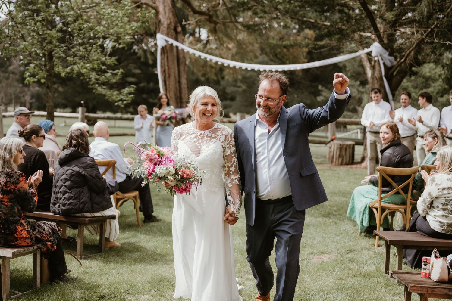 A joyful outdoor wedding ceremony takes place in a lush garden setting with rustic wooden benches and floral arrangements.