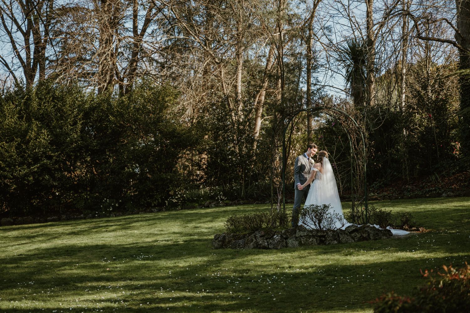 A bride and groom share a romantic moment under a floral arch in a lush green garden surrounded by trees.