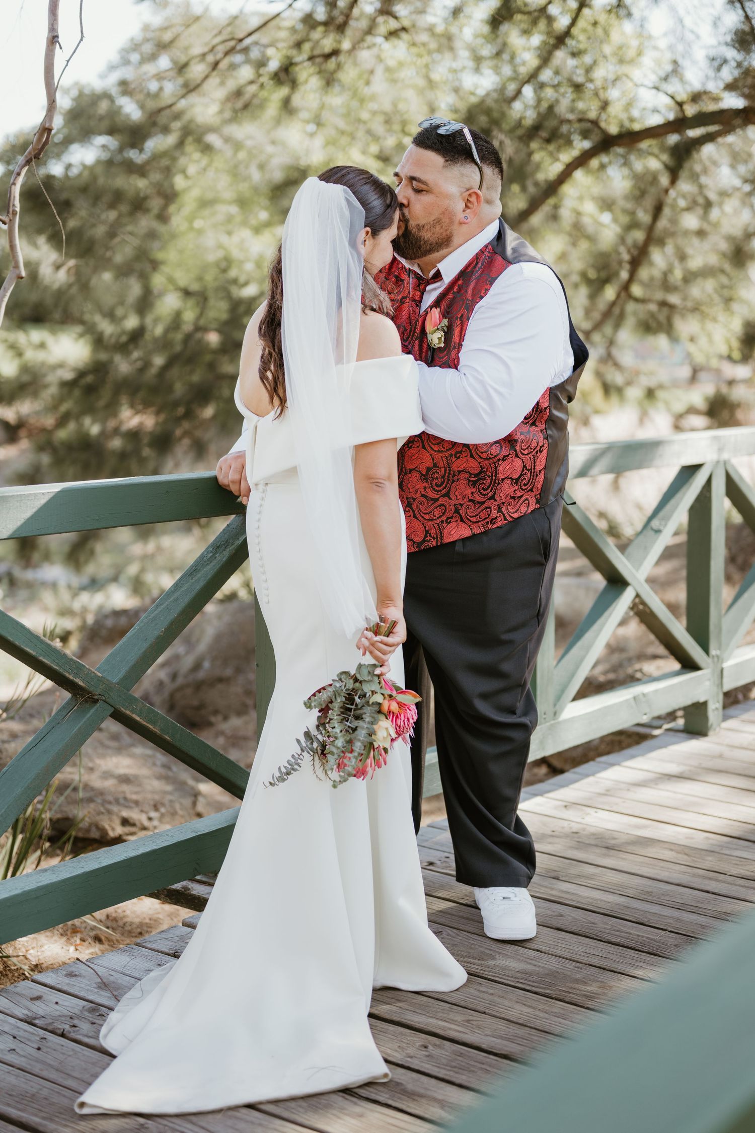 Couple in wedding attire sharing an intimate moment on a wooden bridge surrounded by greenery.