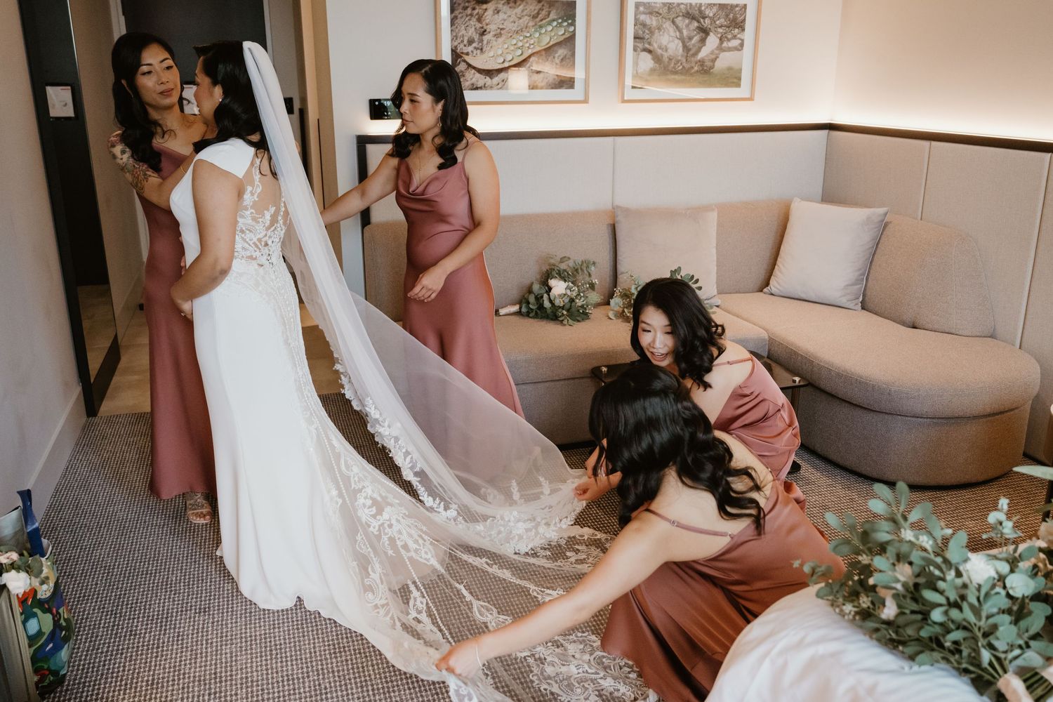 Wedding party getting ready in elegant hotel room with bridesmaids in dusty rose dresses and white decor.