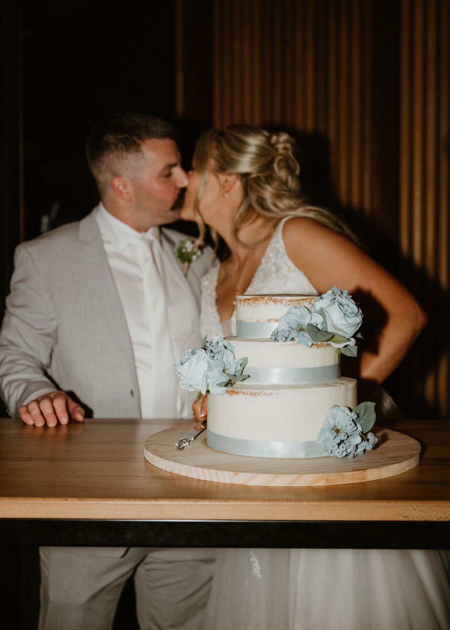 Bride and groom kissing behind a three-tier white wedding cake adorned with blue roses on a wooden table.