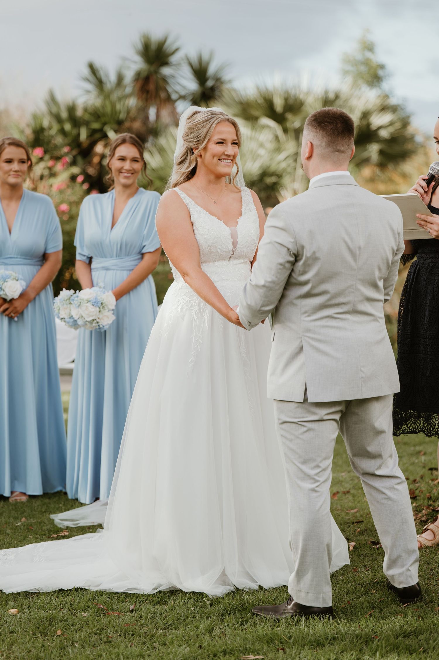 Bride smiling at groom during outdoor wedding ceremony with bridesmaids in light blue dresses in background.