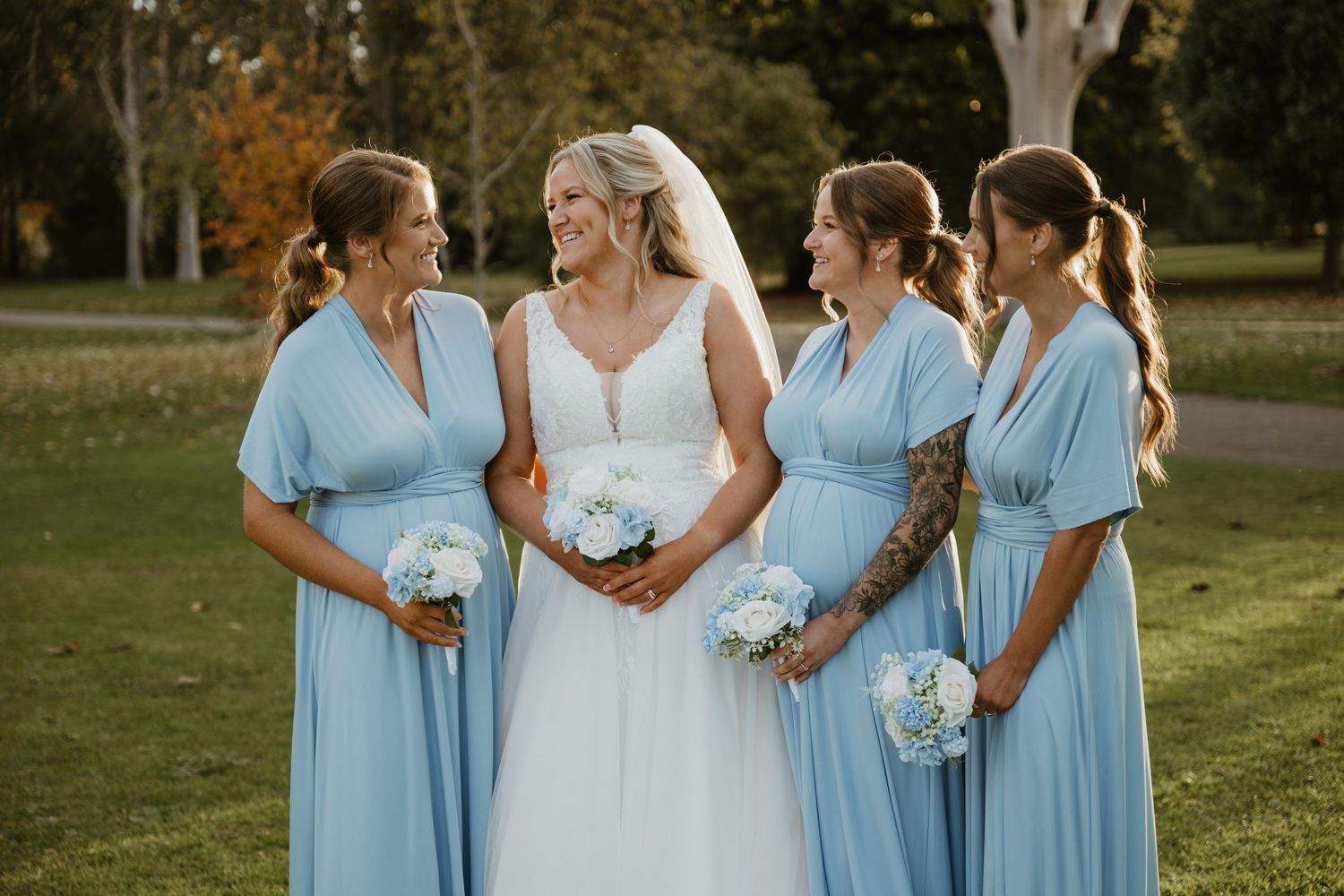 Bride in white gown with three bridesmaids in light blue dresses holding white bouquets outdoors.