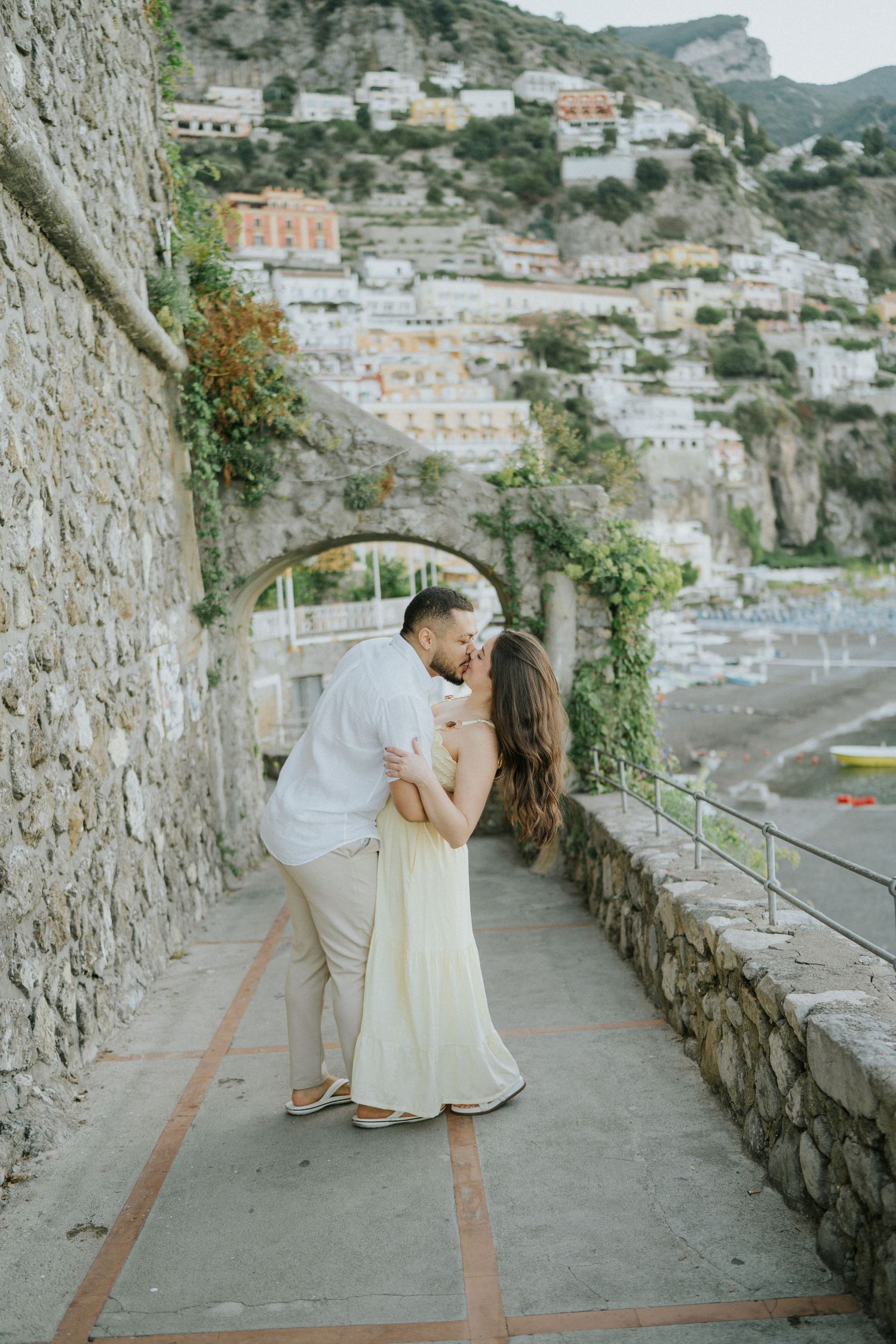 Two people share a romantic dance on a stone pathway with Mediterranean coastal views.