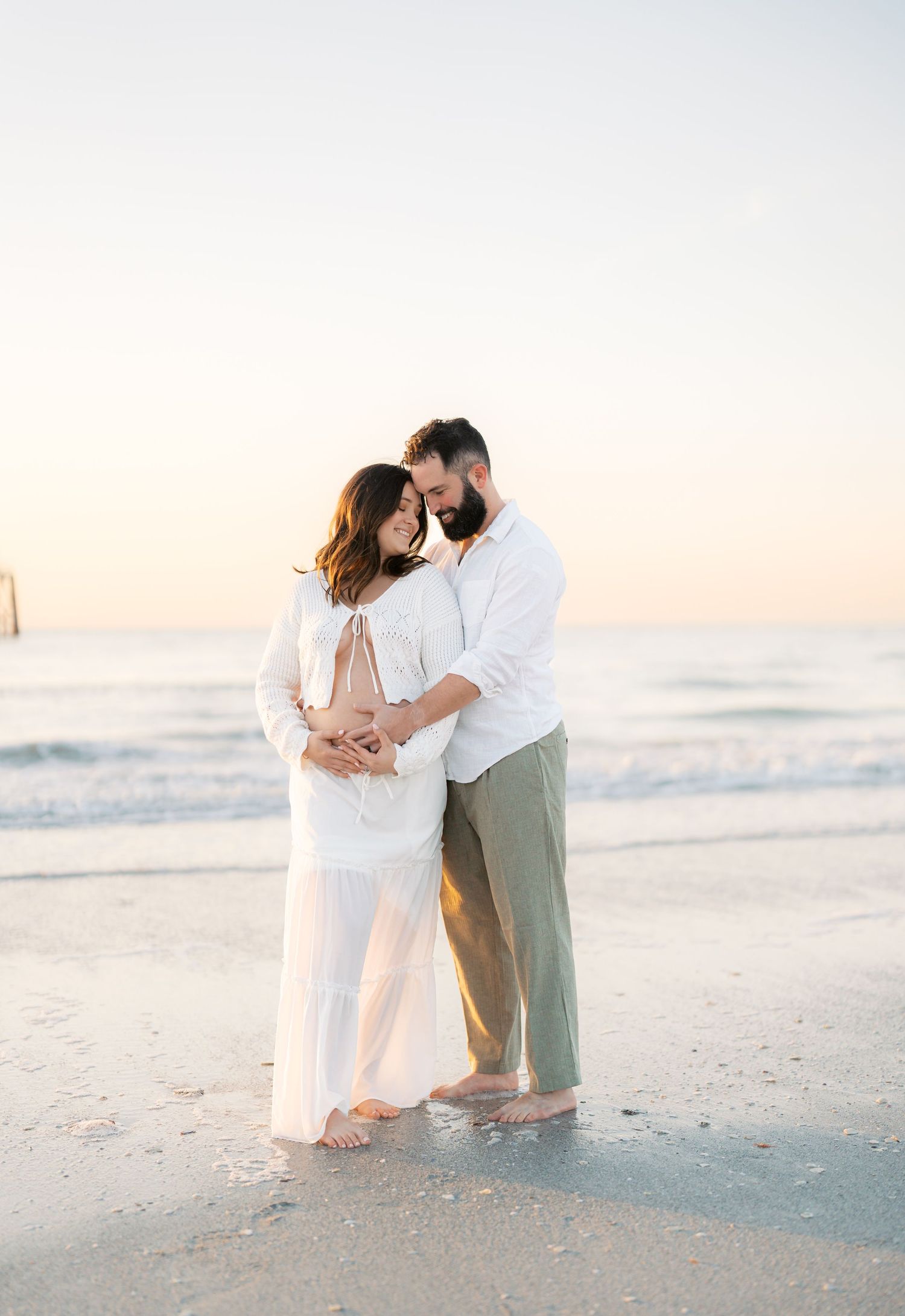 Couple embracing on beach at sunset, woman in white outfit showing baby bump, man in white shirt and olive pants, barefoot.