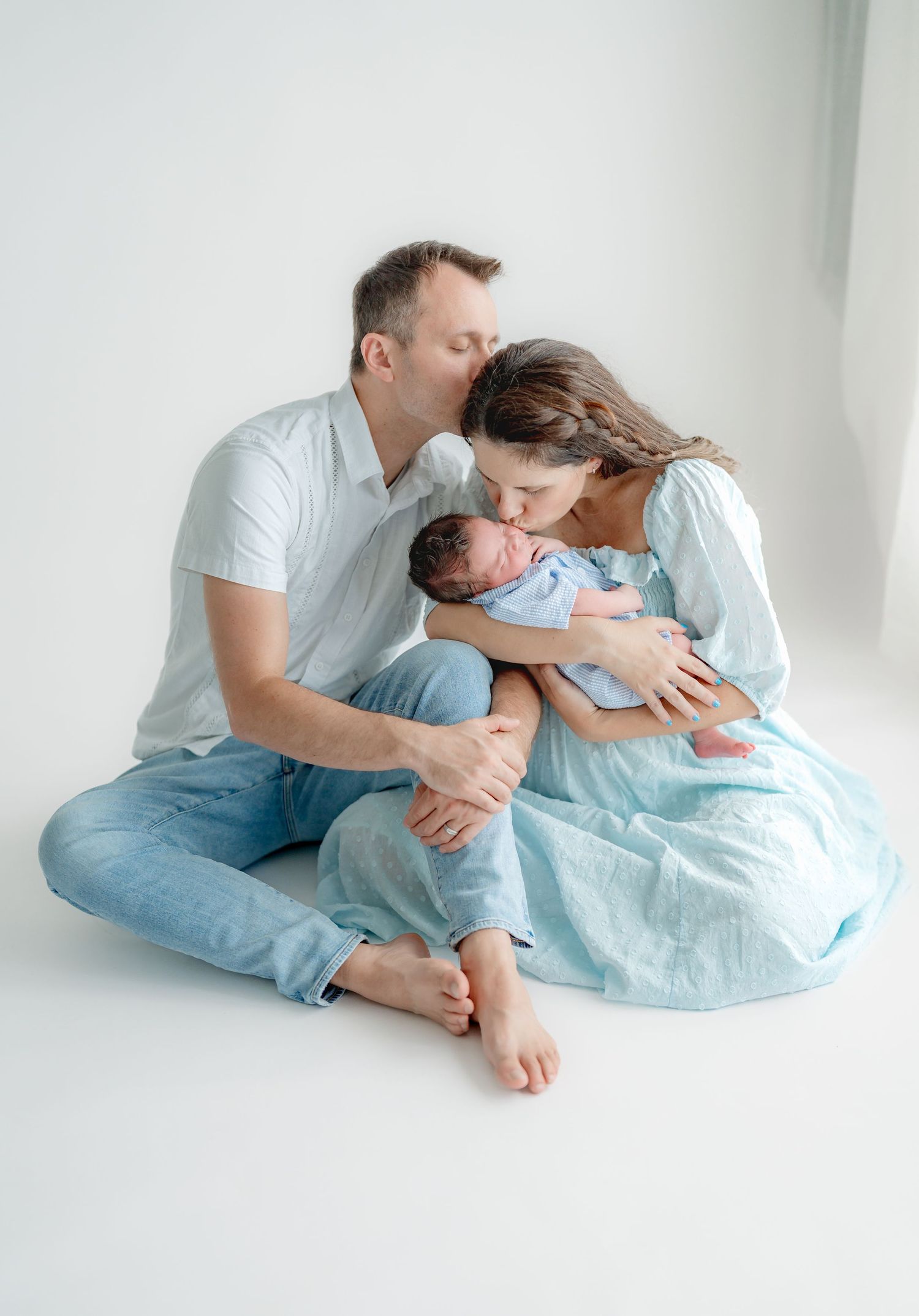 Family lying together on blue fabric in soft white studio setting for intimate lifestyle portraits.