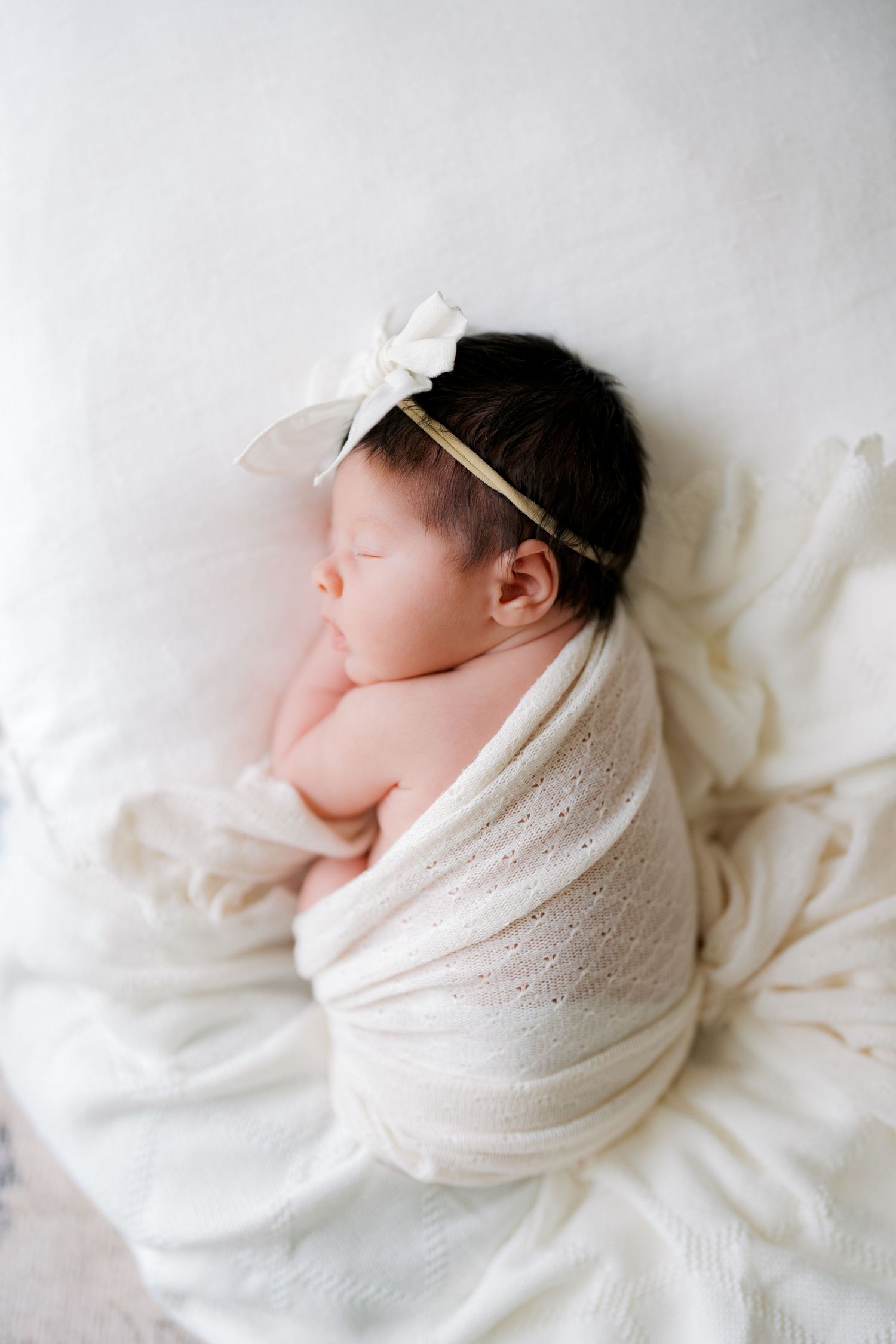 Sleeping newborn baby girl swaddled in cream knit wrap with white bow headband on soft white blanket.