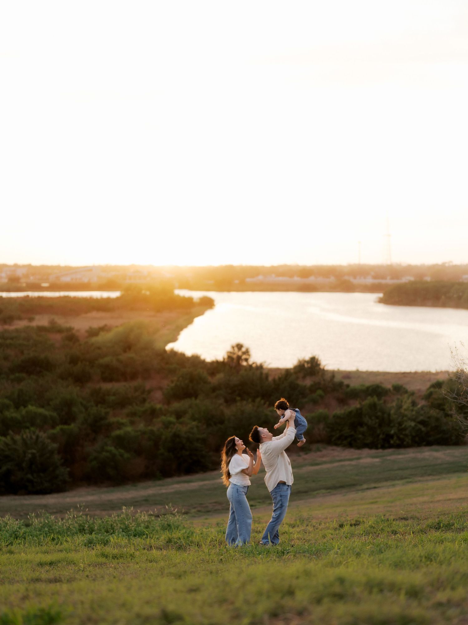 Family of three playing at golden hour on a grassy hill overlooking a scenic lake at sunset.