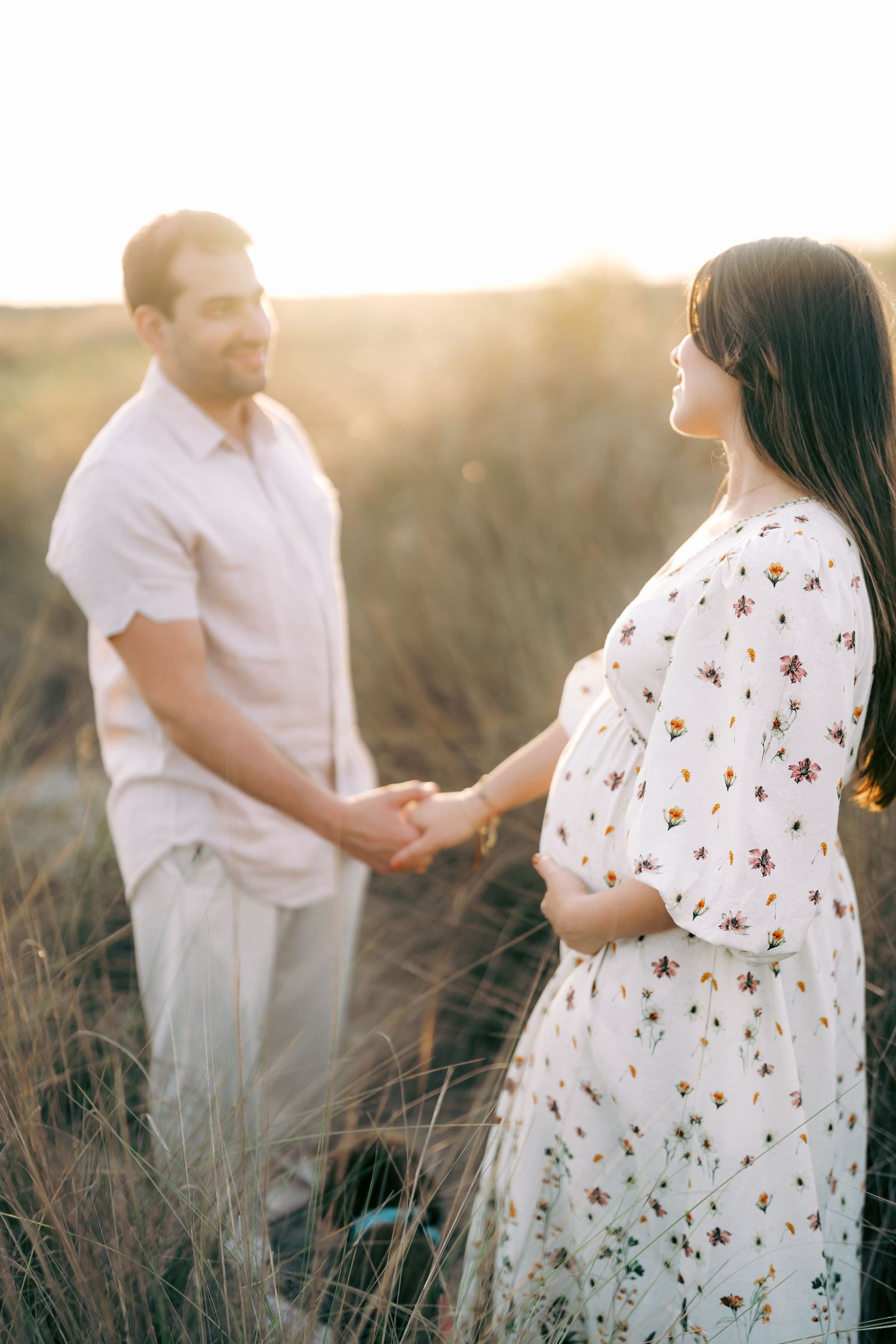 A couple shares a tender moment in a sunlit field wearing white and floral outfits during a romantic photoshoot.