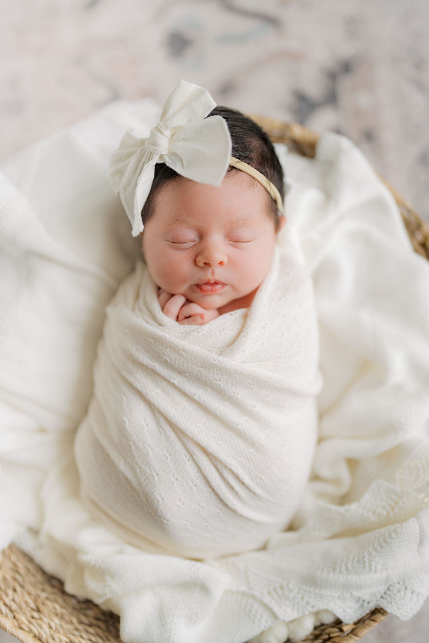 Sleeping newborn baby girl swaddled in white wrap with bow headband, resting peacefully in a wicker basket.