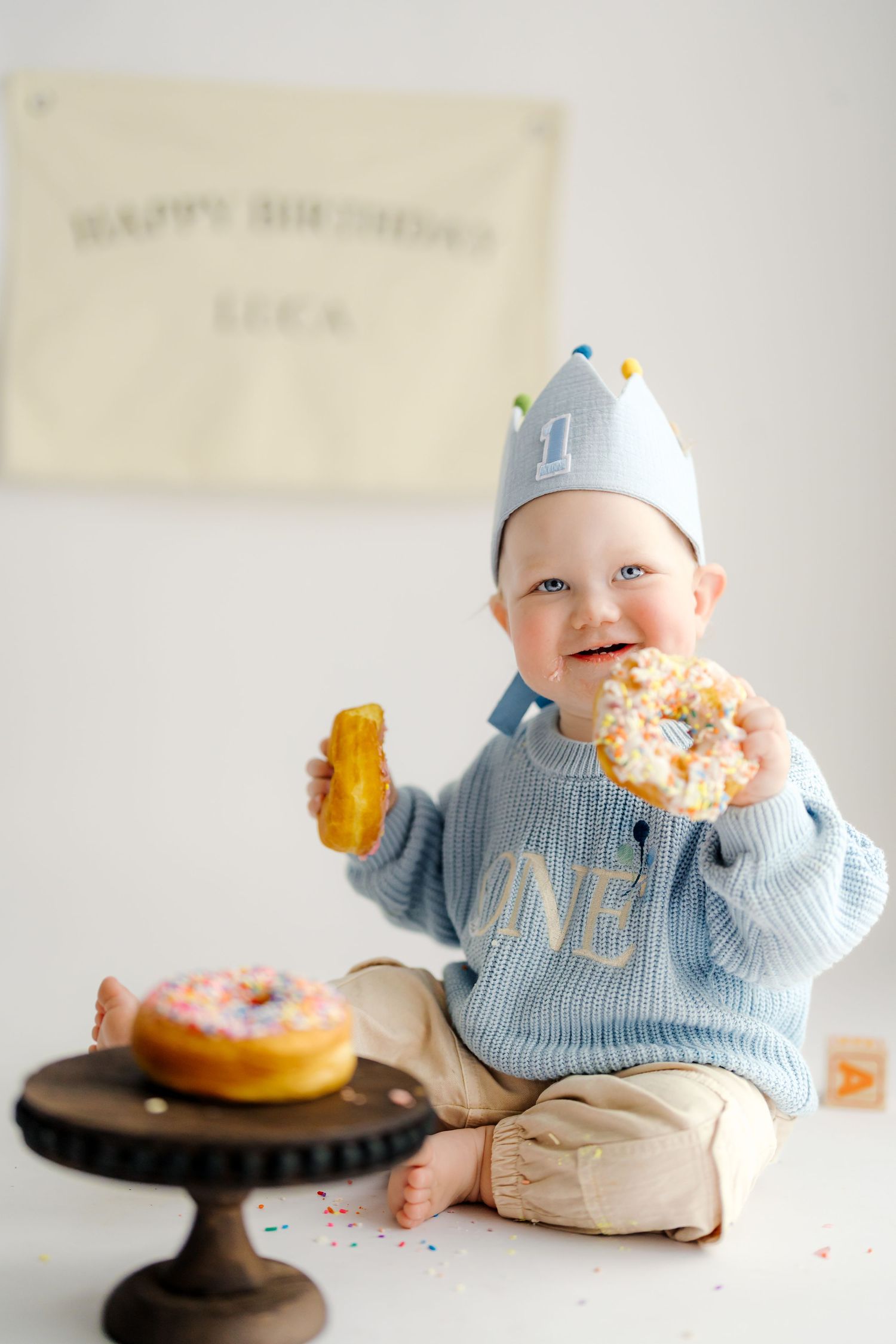 Baby in blue sweater enjoys birthday cake smash session with pink frosted donut on wooden pedestal.