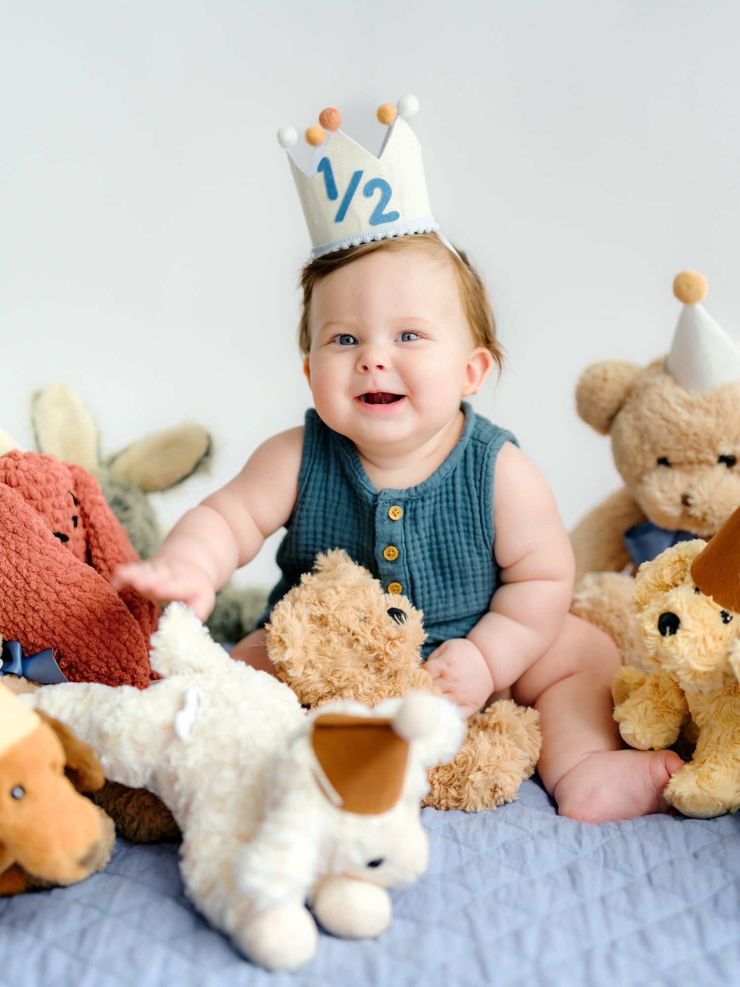 A baby in a blue knit vest sits on a white blanket surrounded by plush teddy bears and stuffed animals.