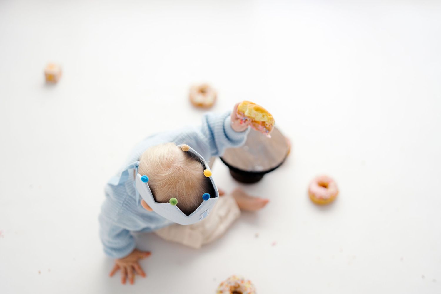 Baby in blue onesie reaching for glazed donuts scattered on white background.