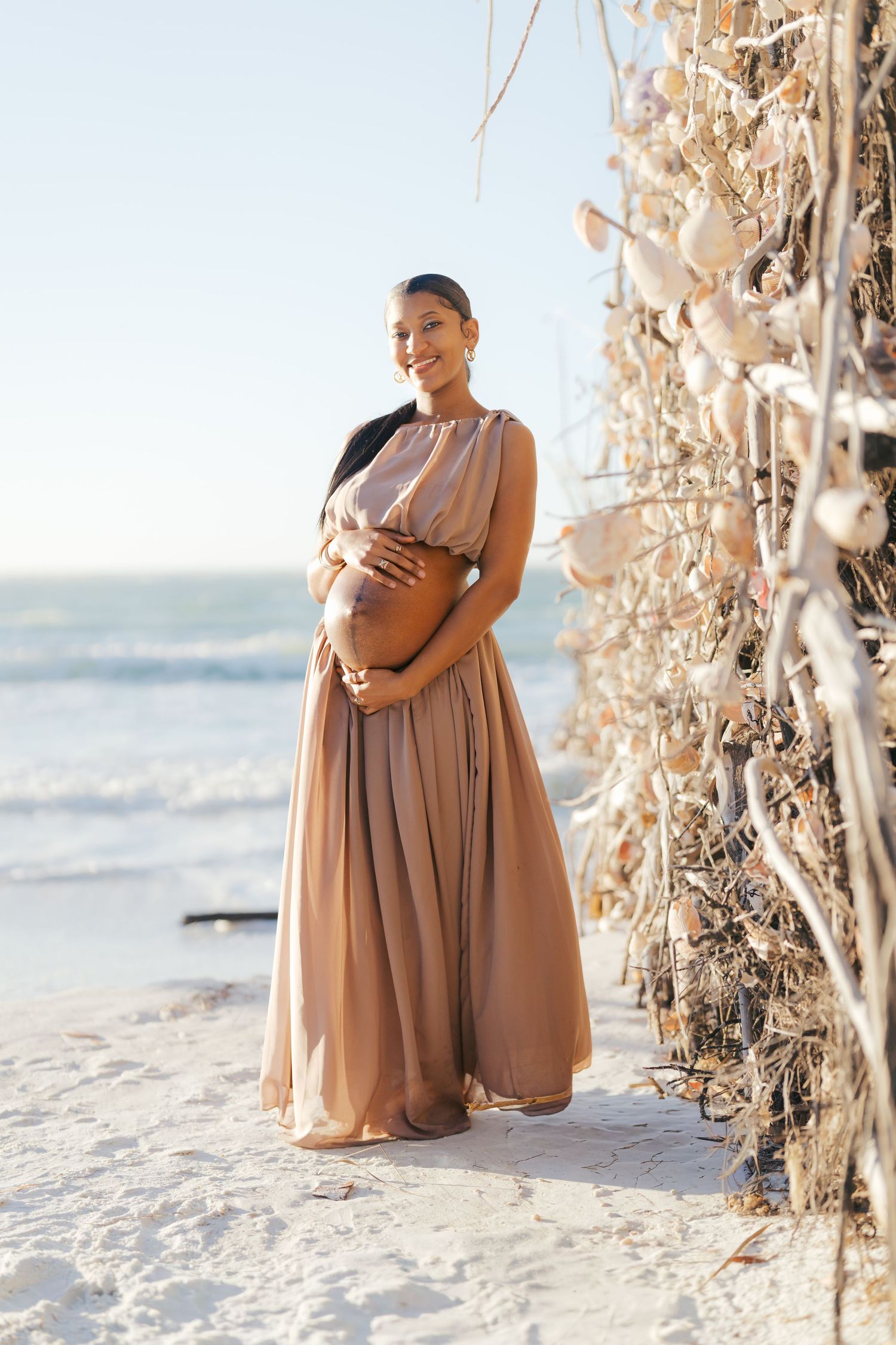 Pregnant woman in beige maxi dress cradling baby bump on sandy beach near driftwood decorated with shells.