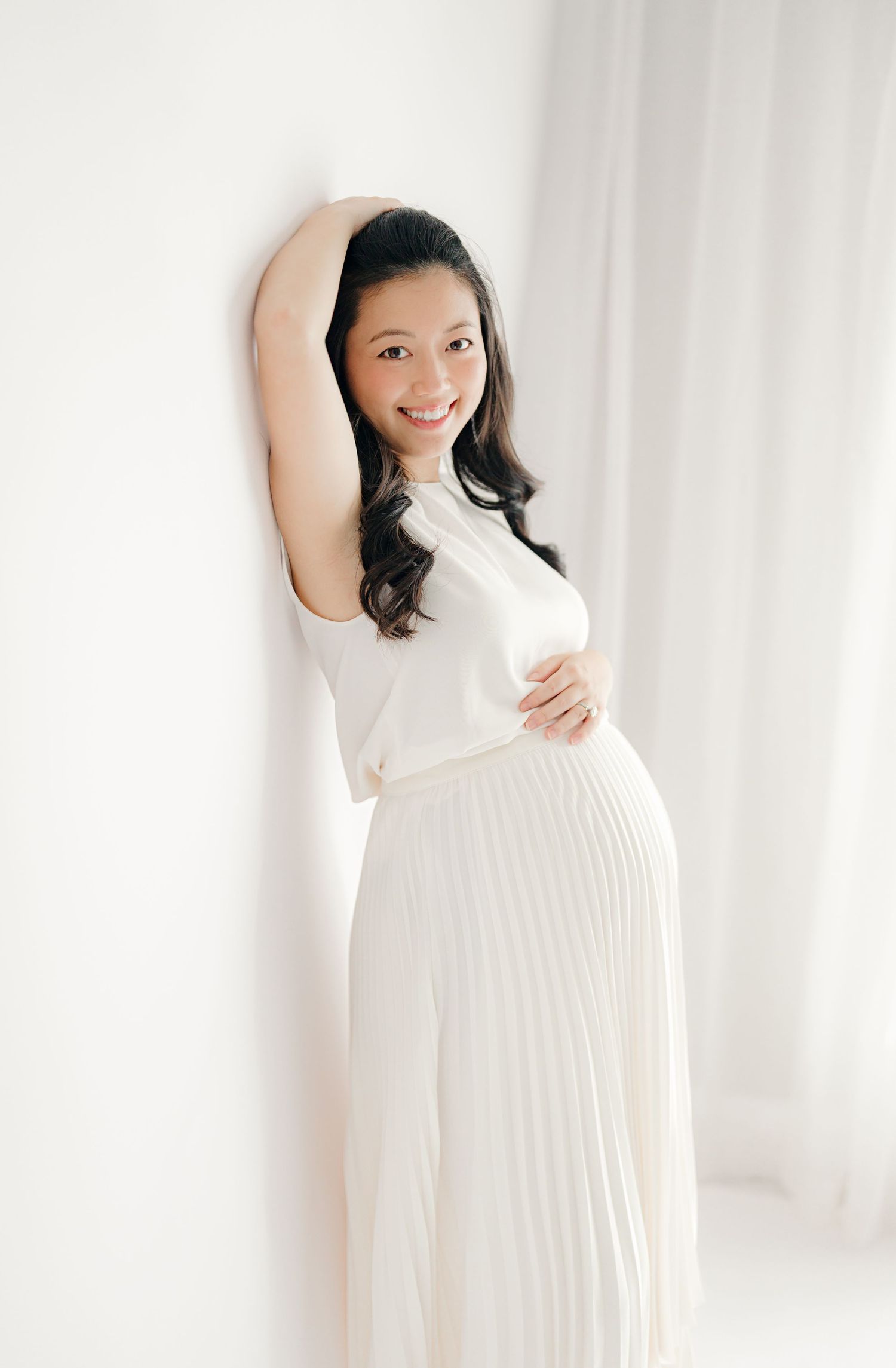 Minimalist studio maternity photos with woman in white pleated dress against white backdrop.