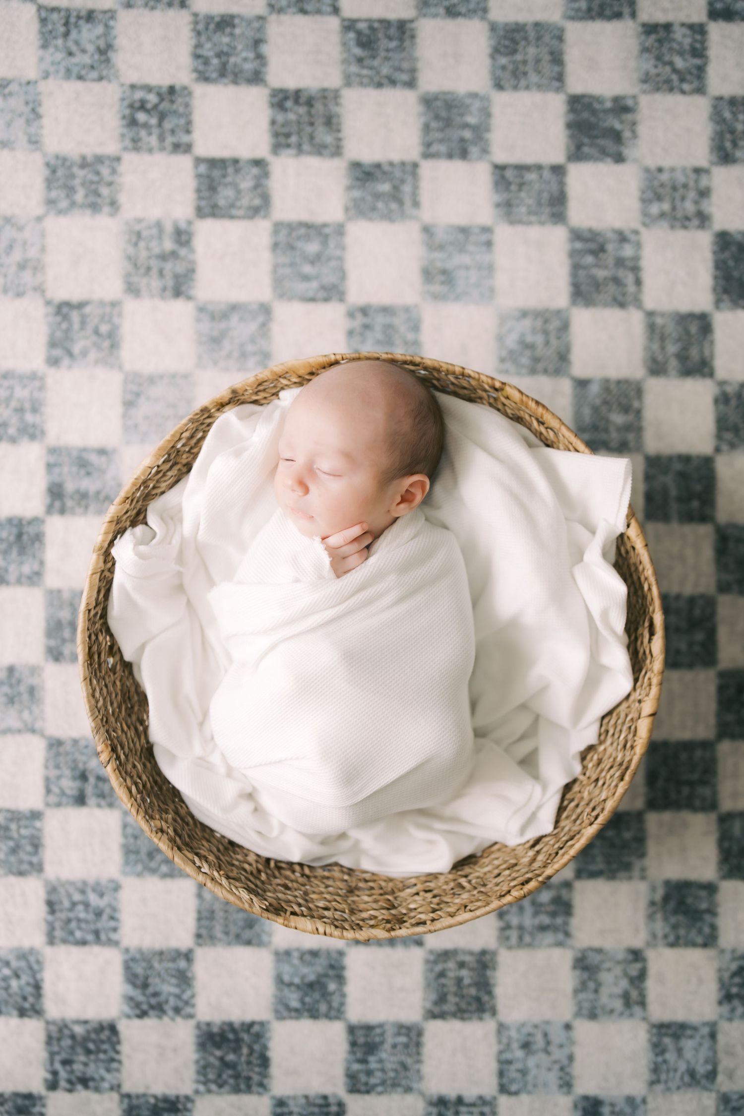 Sleeping newborn wrapped in white swaddle lies in woven basket on checkered floor.