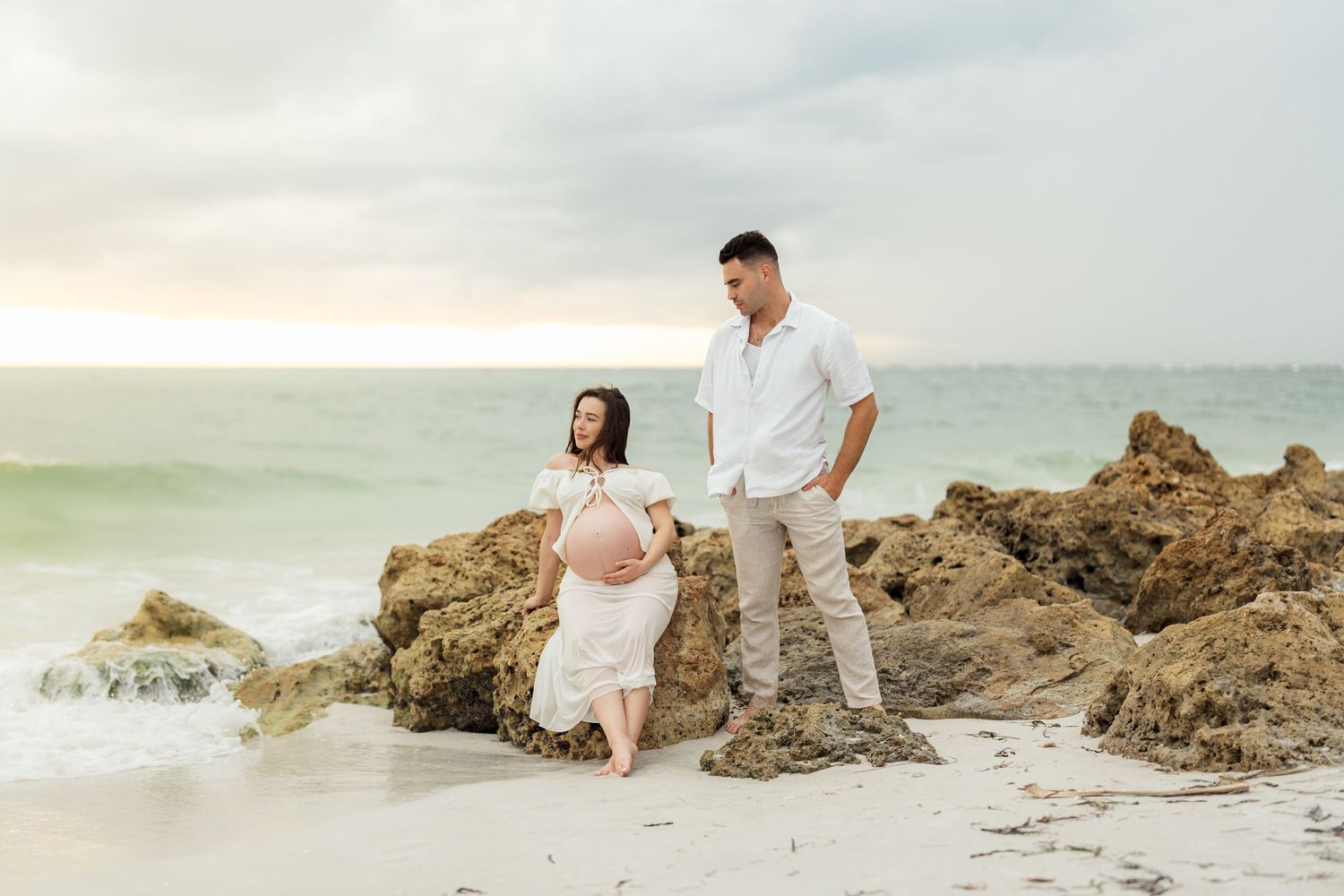 Couple standing on rocky beach during sunset for maternity photo session.