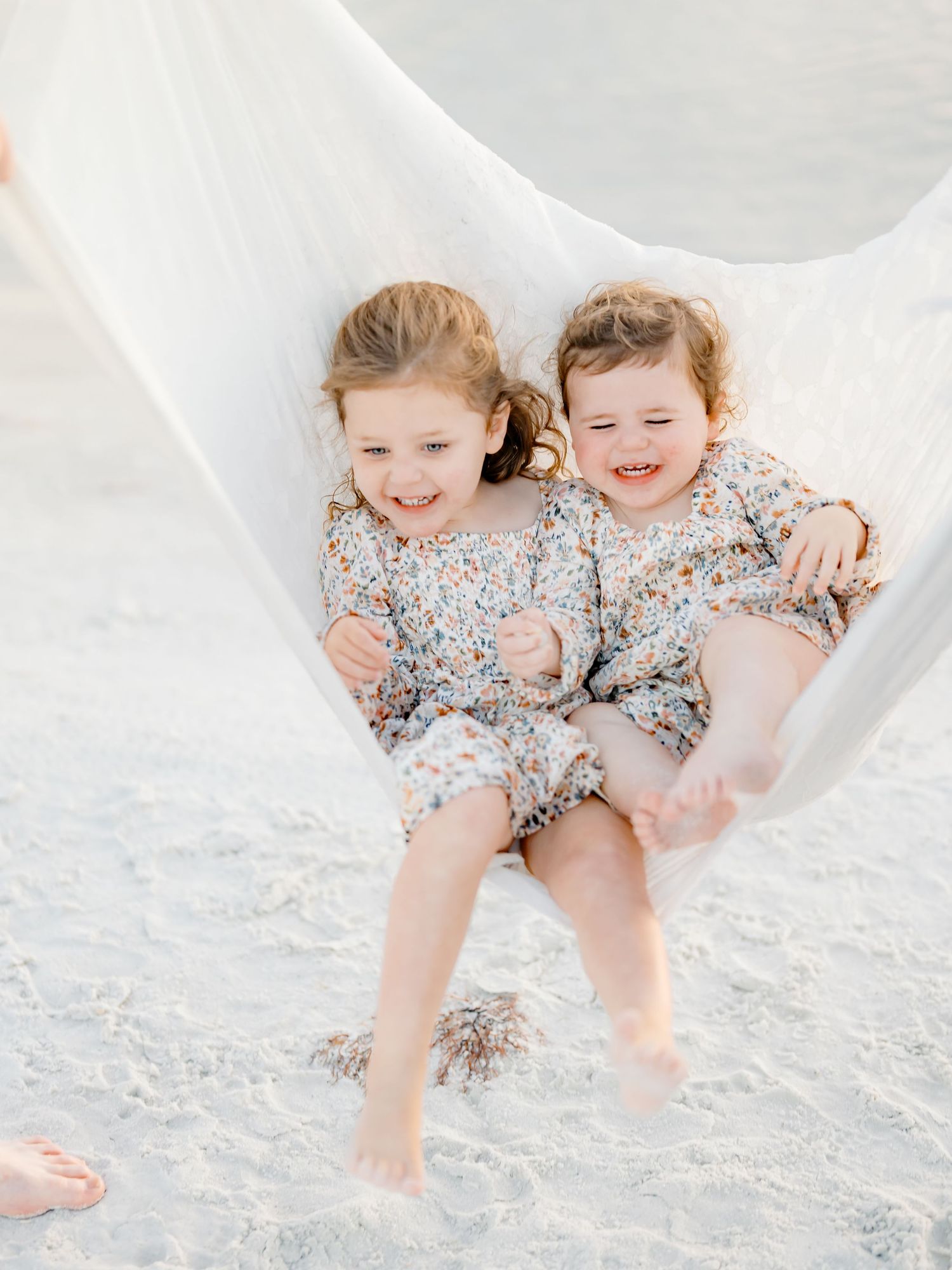 Two children in matching floral outfits playing and laughing together on a white sandy beach.