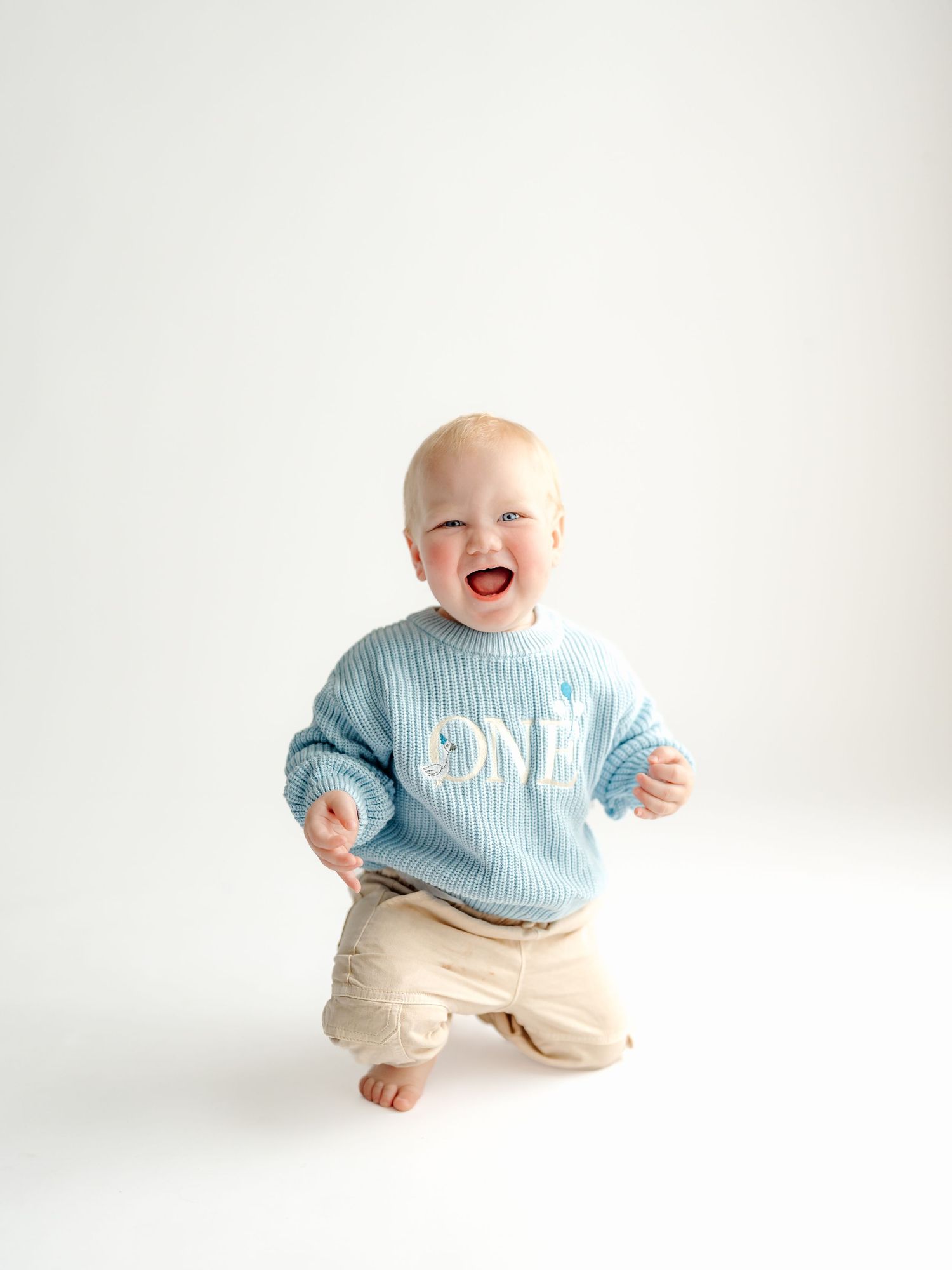 A baby wearing a light blue striped 'ONE' shirt and khaki pants sits and laughs joyfully against a white background.