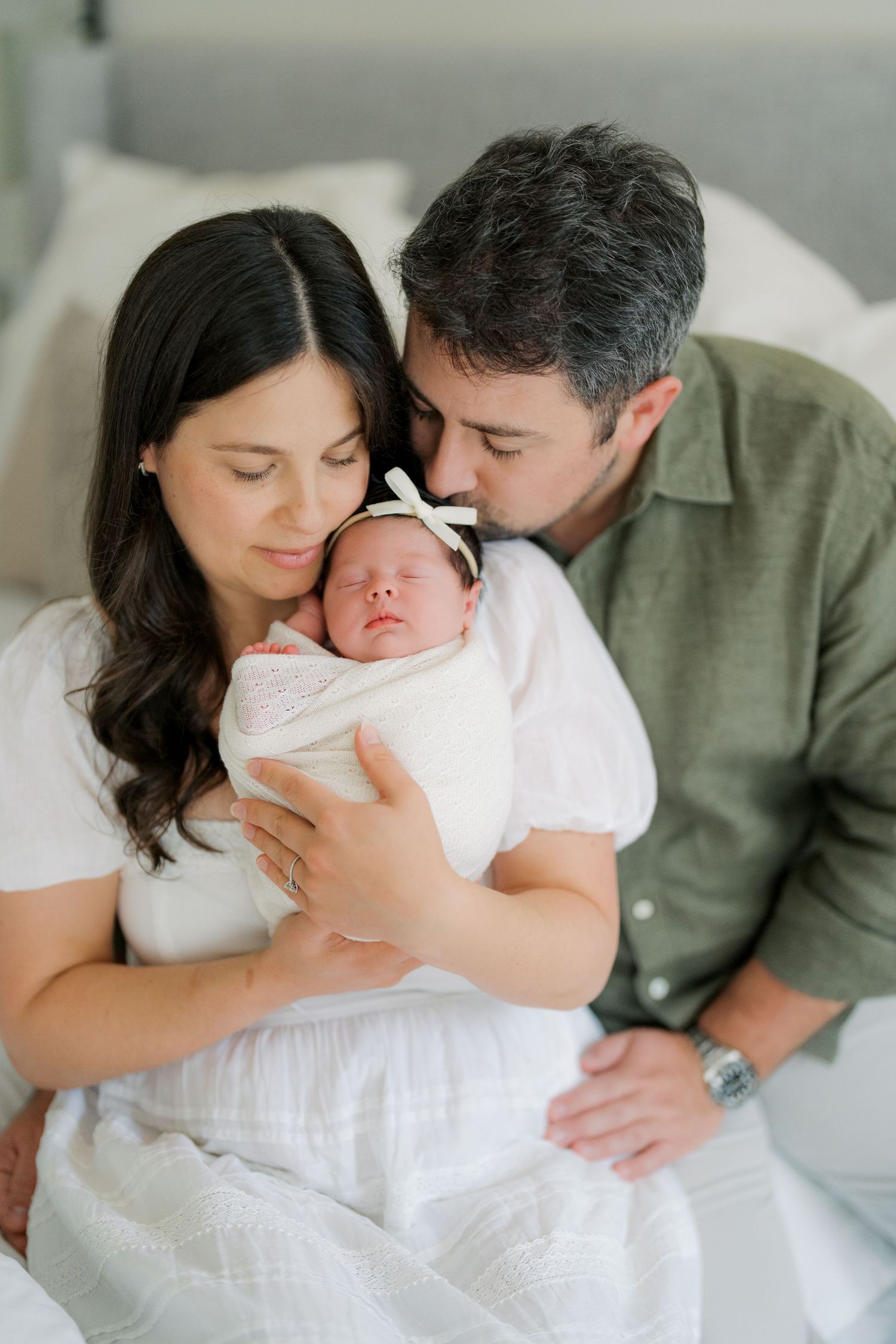 New parents tenderly hold their swaddled newborn baby girl with white bow headband during an intimate newborn photo session.