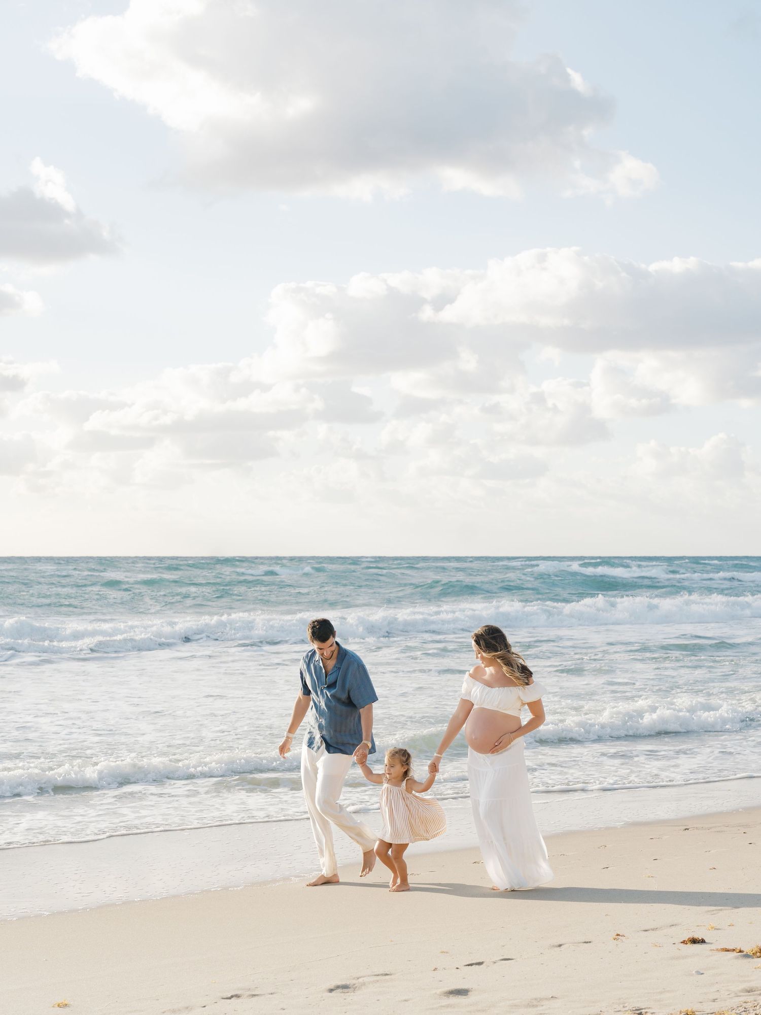Pregnant woman in white dress walks on beach with partner and toddler, ocean waves behind them.