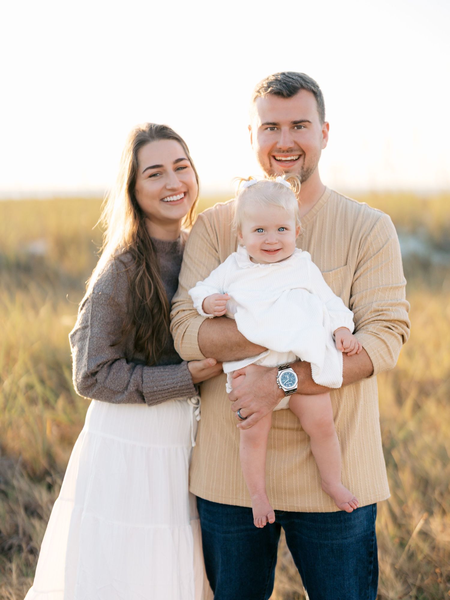 A family of three poses together in a golden field at sunset wearing neutral colored clothing.