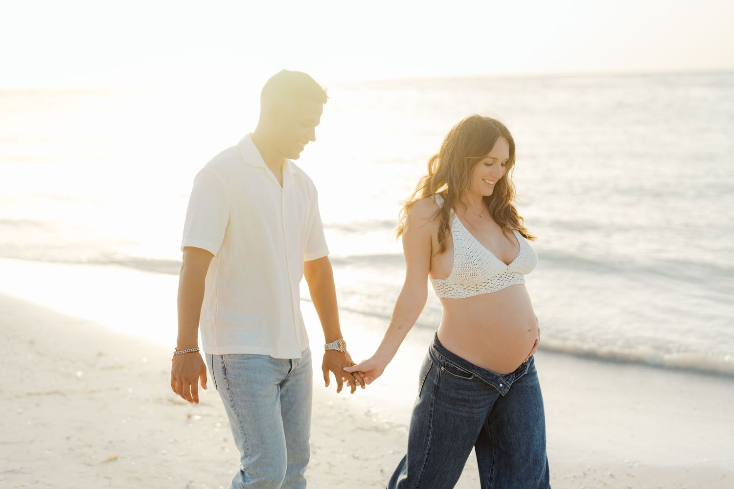 Expectant couple walking hand in hand on beach at sunset.