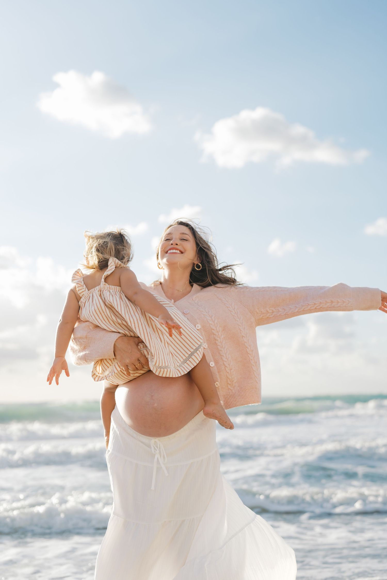 Pregnant woman joyfully lifting toddler at beach, ocean waves behind them on sunny day.